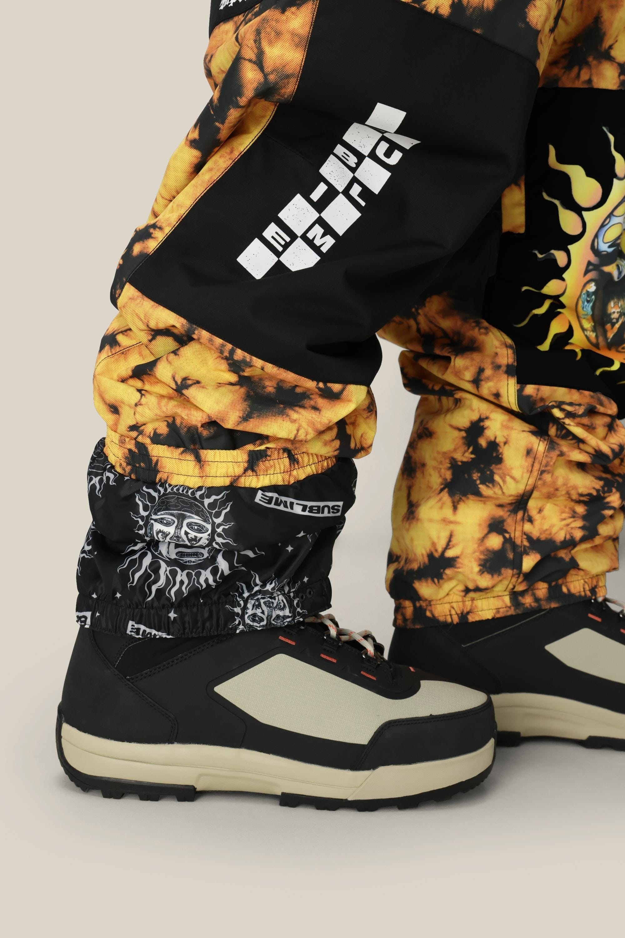 Close-up of a persons lower legs wearing 686 Mens Sublime Shell Pants in yellow and black tie-dye, paired with black gaiters with sun and moon designs, plus black-and-beige outdoor shoes, all set against a neutral background.