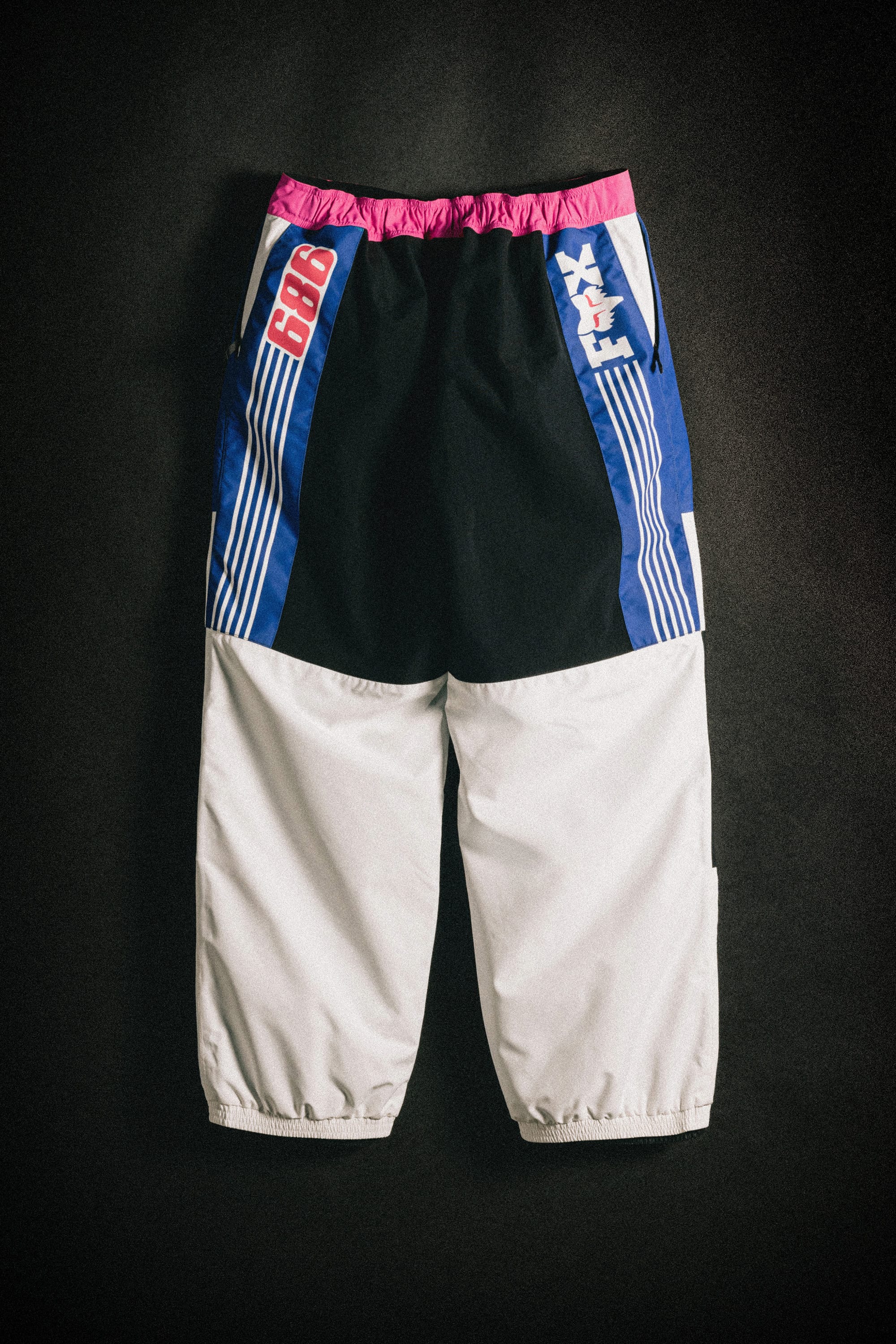 The 686 Men's Fox Racing Shell Pant by 686 features a pink elastic waistband, black upper, blue side panels with white stripes and text, and light gray lower legs, shown against a dark background.