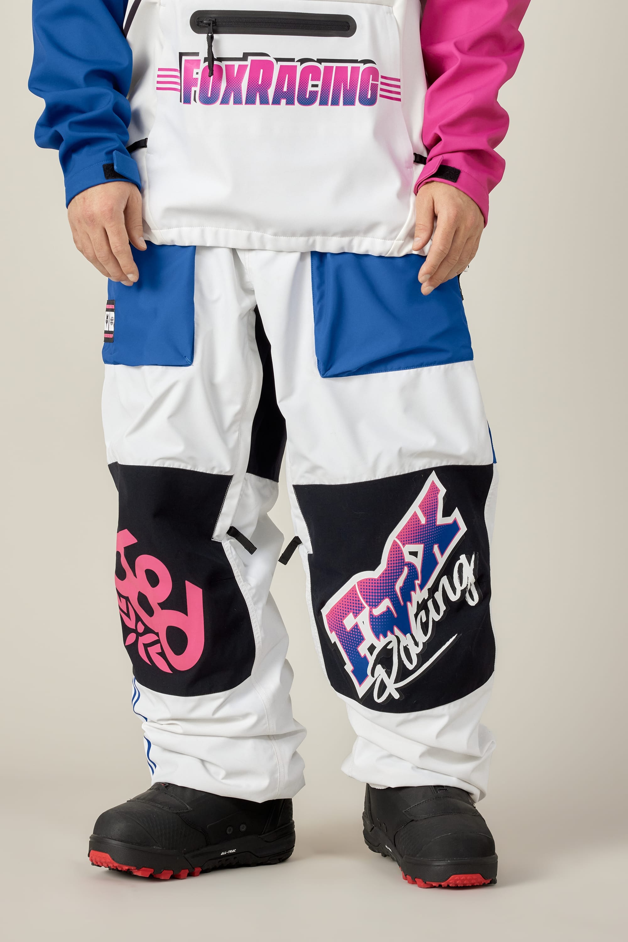A person stands against a plain background wearing the 686 Men's Fox Racing Shell Pant, featuring blue, white, and pink racing gear with large Fox Racing logos on the jacket and pants, paired with black boots.