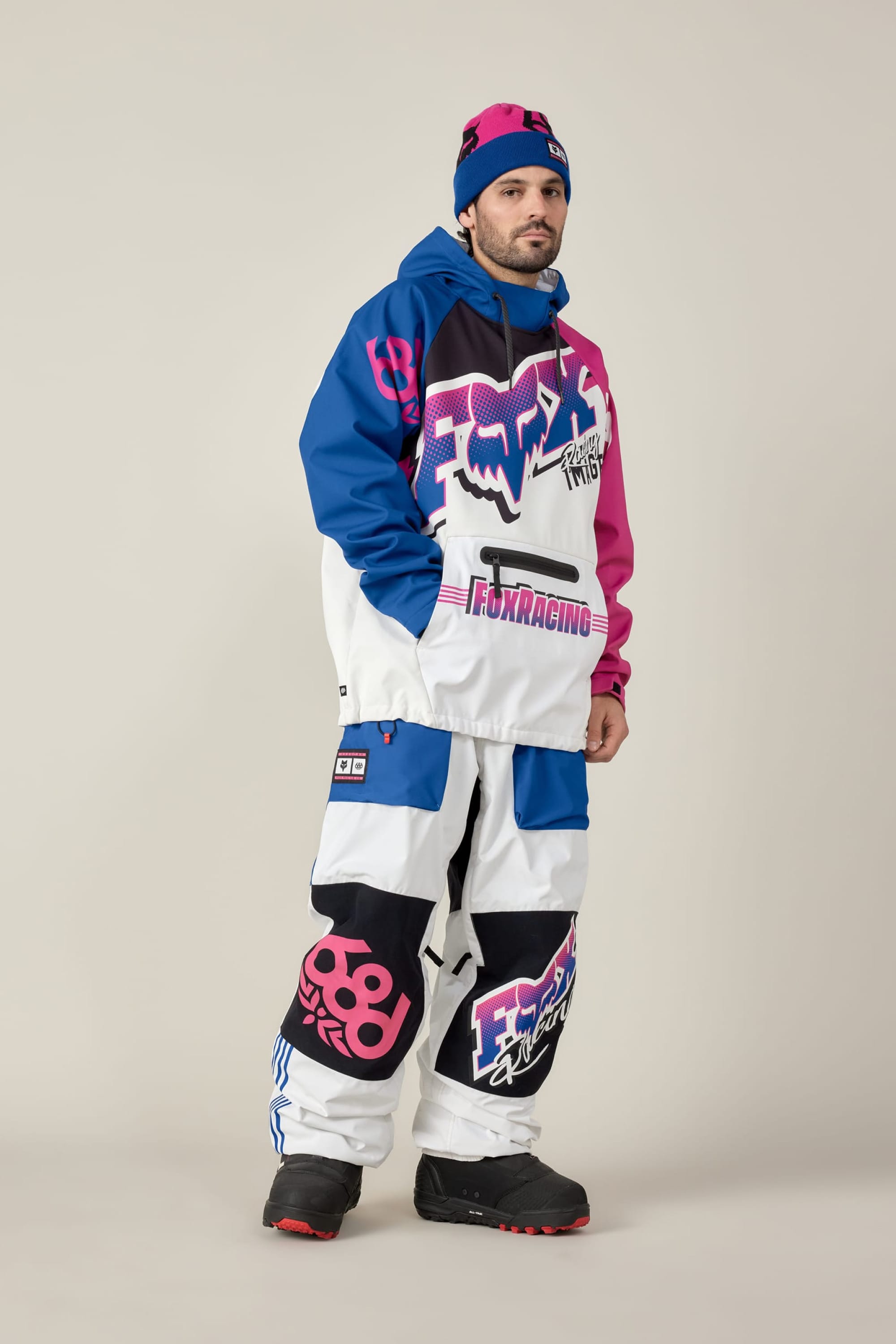 A man stands against a plain light background wearing a bold, colorful winter outfit with a blue, pink, and white Fox Racing hoodie, 686 Men's Fox Racing Shell Pant by 686, a matching beanie, and black snow boots.