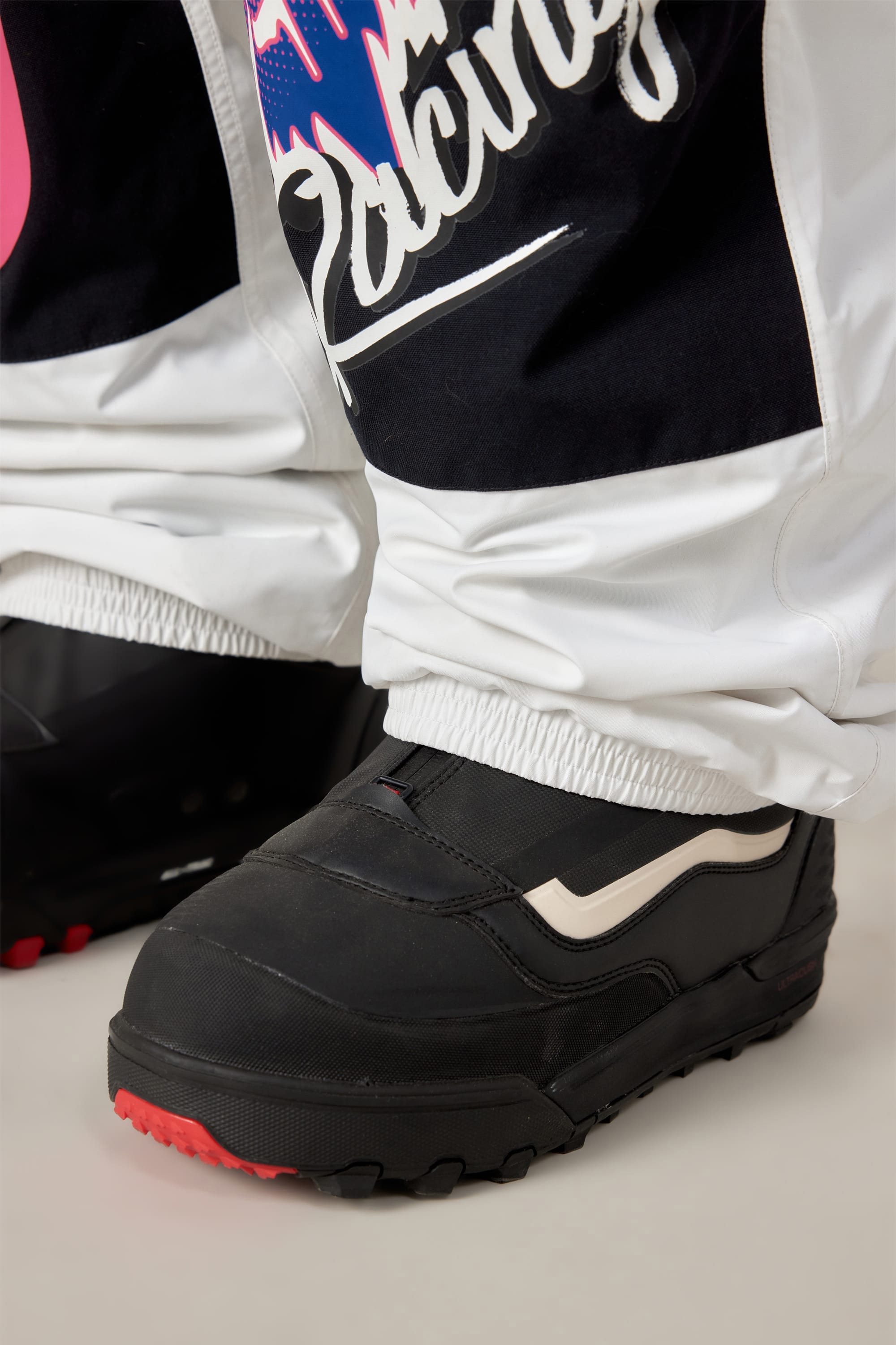 Close-up of a person wearing black snowboarding boots and 686 Men's Fox Racing Shell Pant, featuring a colorful graphic and the word "Racing," by 686.