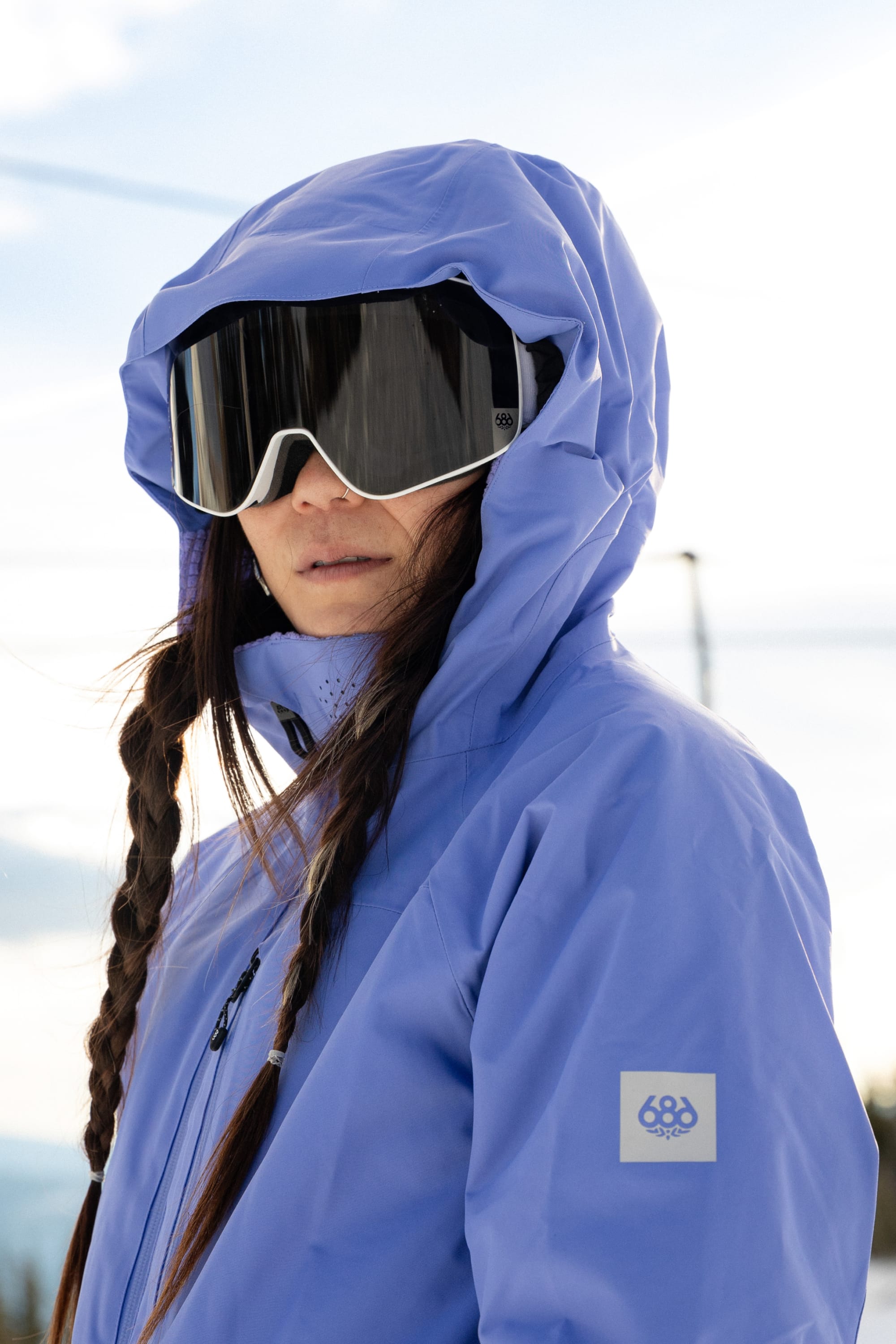 A person wearing the 686 Women’s Hydra™ Thermagraph® Jacket, ski goggles, and sporting braids stands outdoors on a snowy day.
