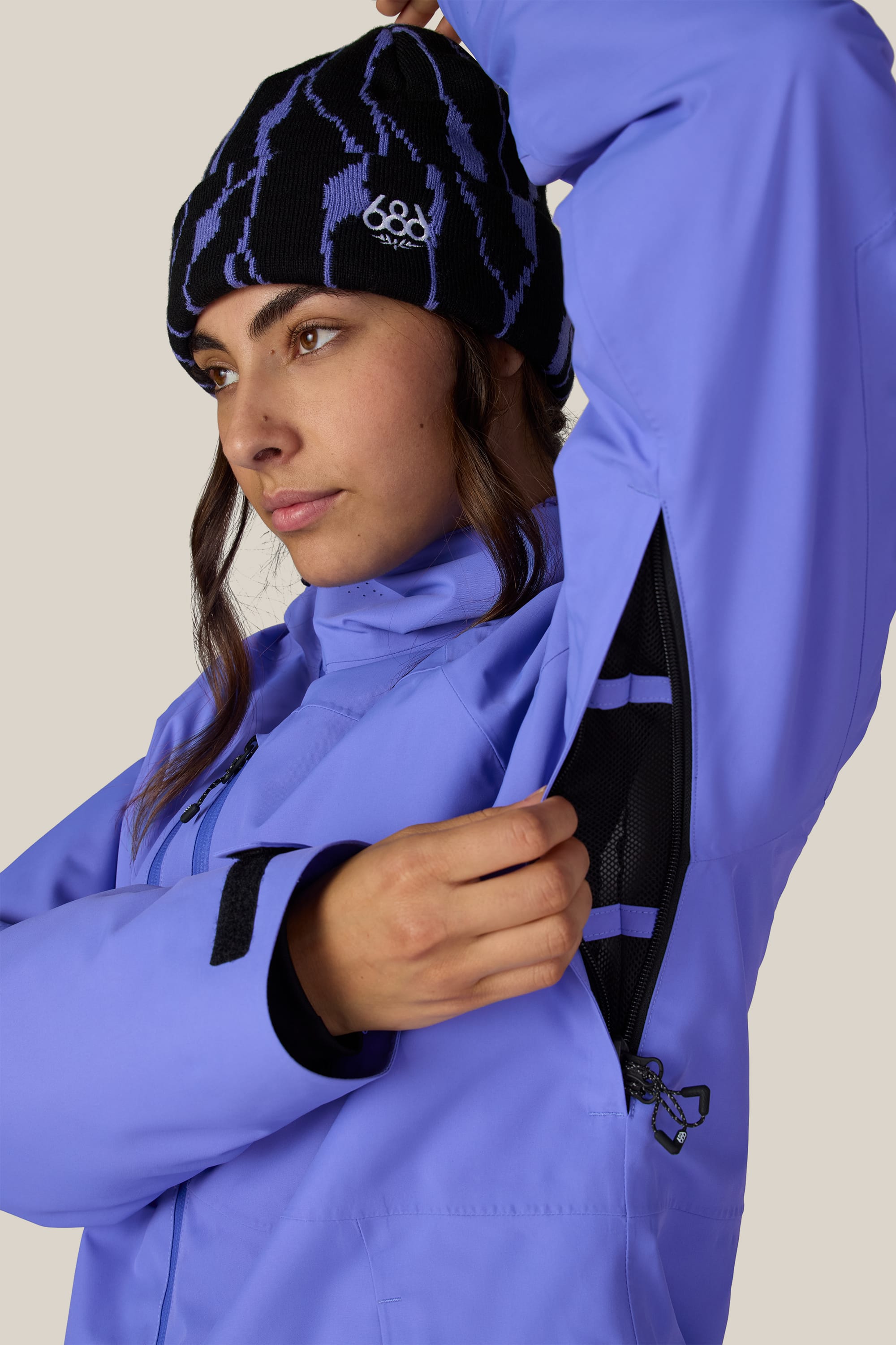 A woman wearing the 686 Women’s Hydra™ Thermagraph® Jacket in purple and a patterned beanie unzips the underarm ventilation zipper, revealing the black mesh lining inside. She gazes to the side with a neutral expression against a plain background.