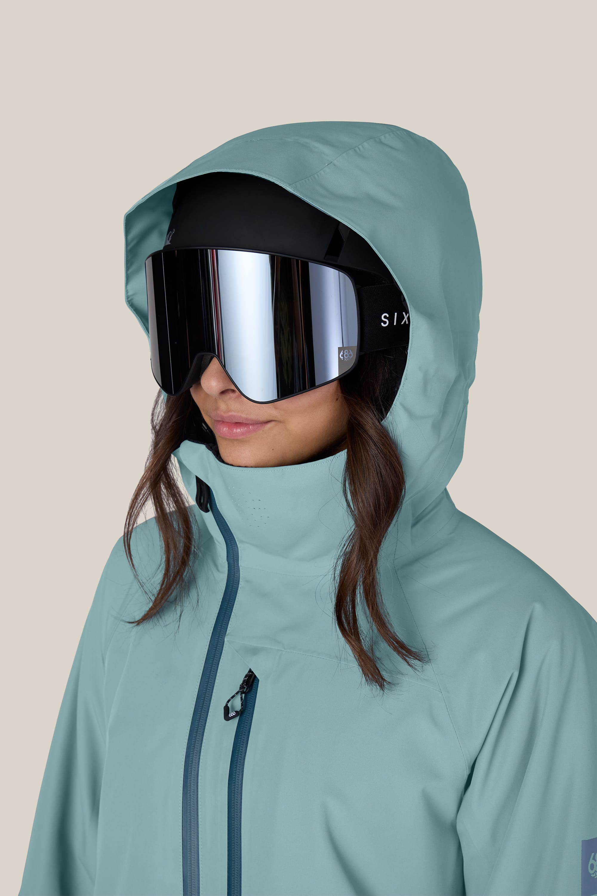 A woman wearing the 686 Women’s Hydra™ Thermagraph® Jacket in teal and large reflective ski goggles stands against a plain beige background.