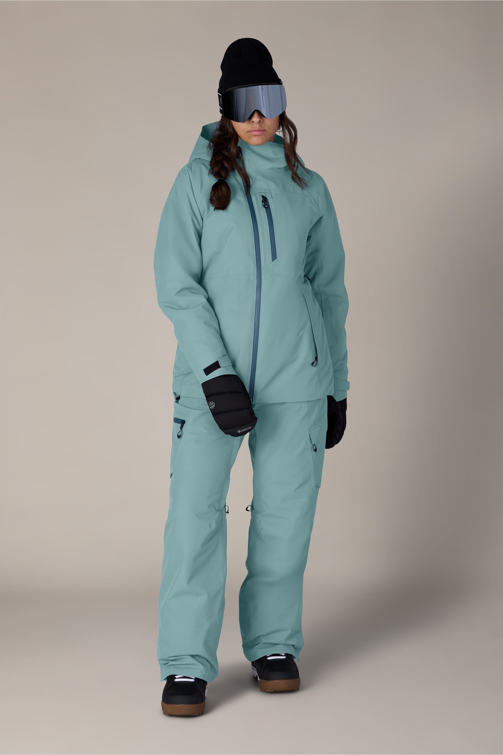 A person wearing the 686 Women’s Hydra™ Thermagraph® Jacket by 686 in light blue, paired with matching pants, black gloves and boots, a black beanie, and large reflective ski goggles, stands against a plain light background.