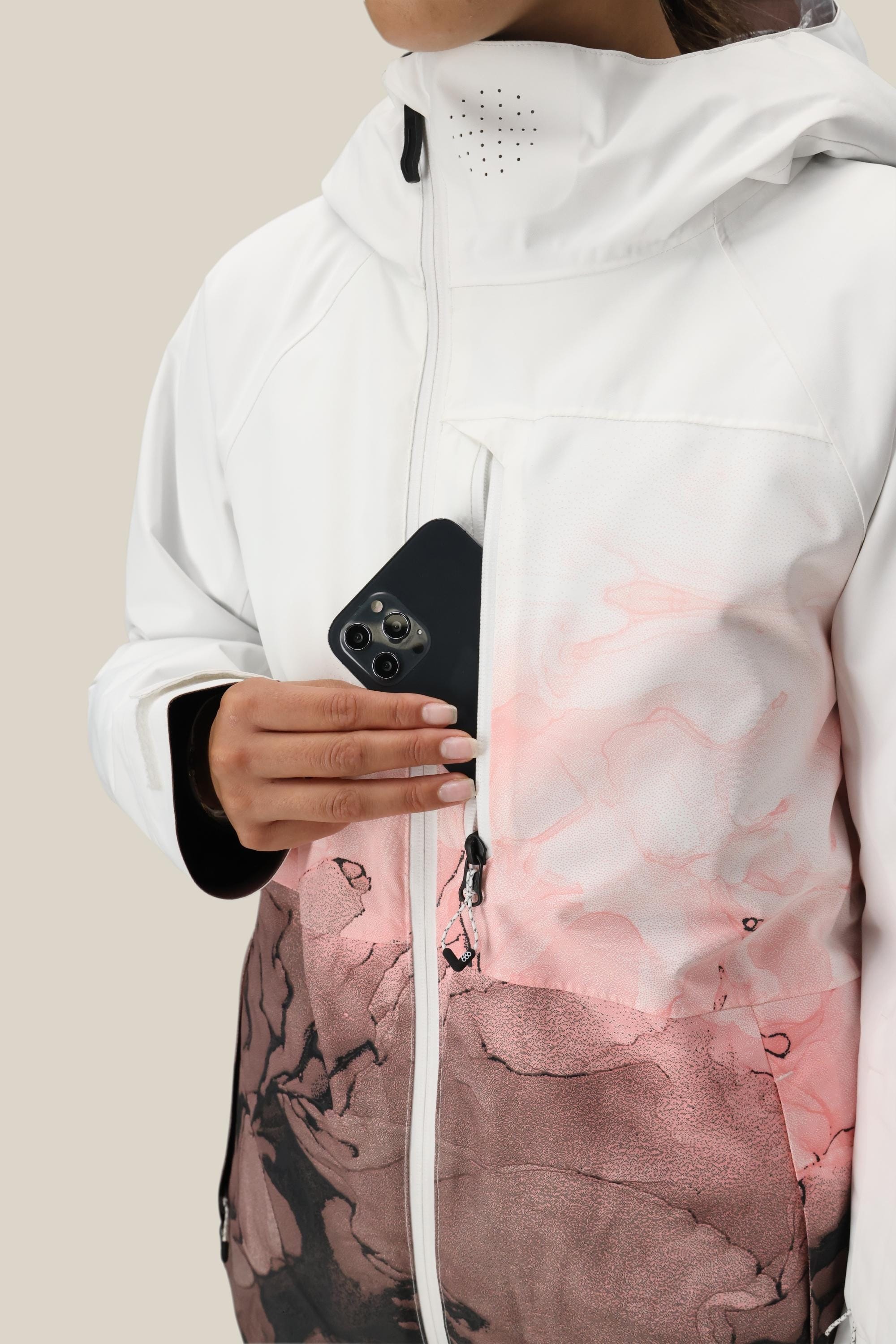 A person in a white and pink 686 Women's Hydra™ Thermagraph® Jacket with hood holds a black smartphone halfway inside the zippered chest pocket against a plain, light background.