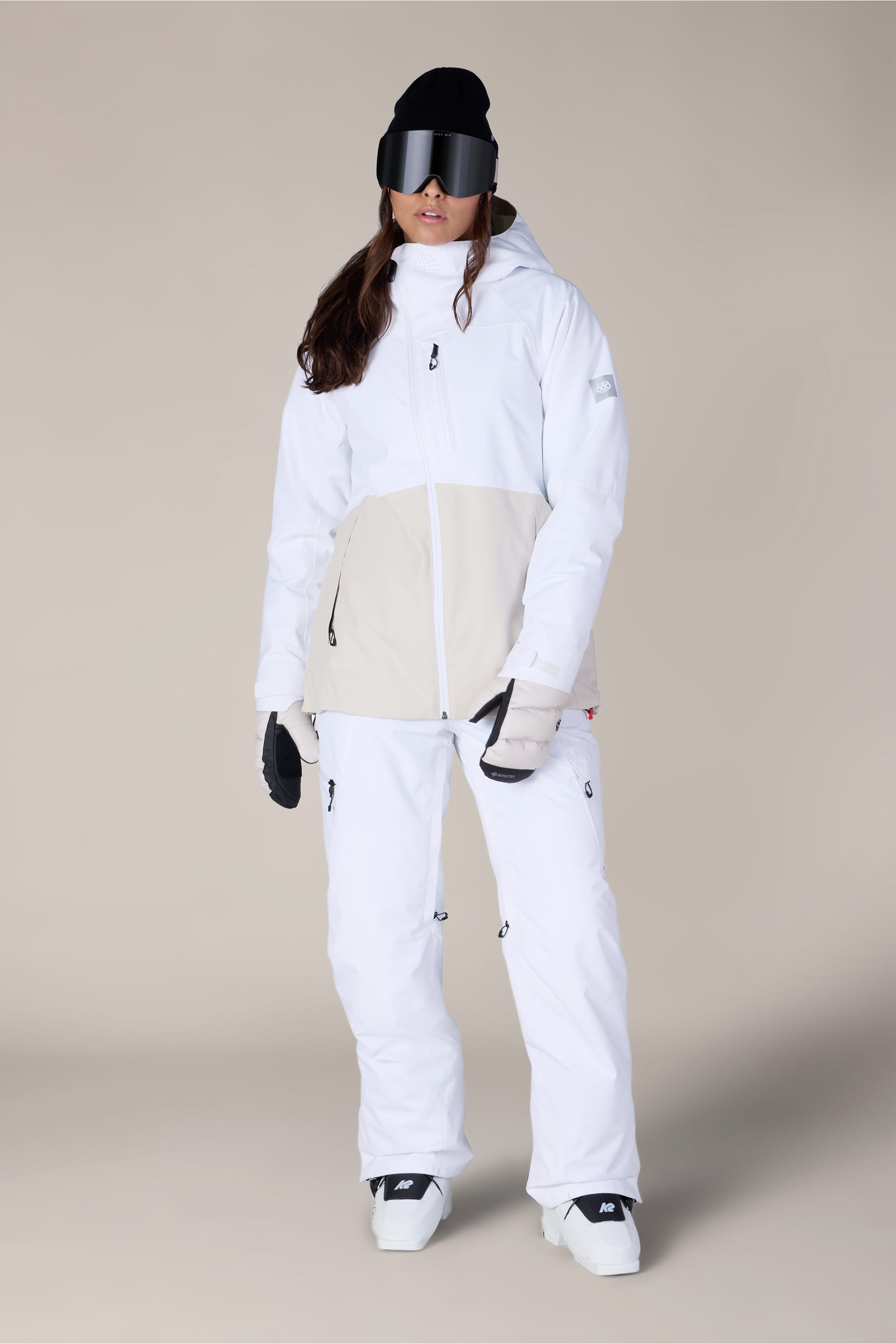 A person wearing the 686 Women’s Hydra™ Thermagraph® Jacket by 686 in white and beige, paired with matching white snow pants, black gloves, and black snow goggles stands facing forward against a plain beige background.