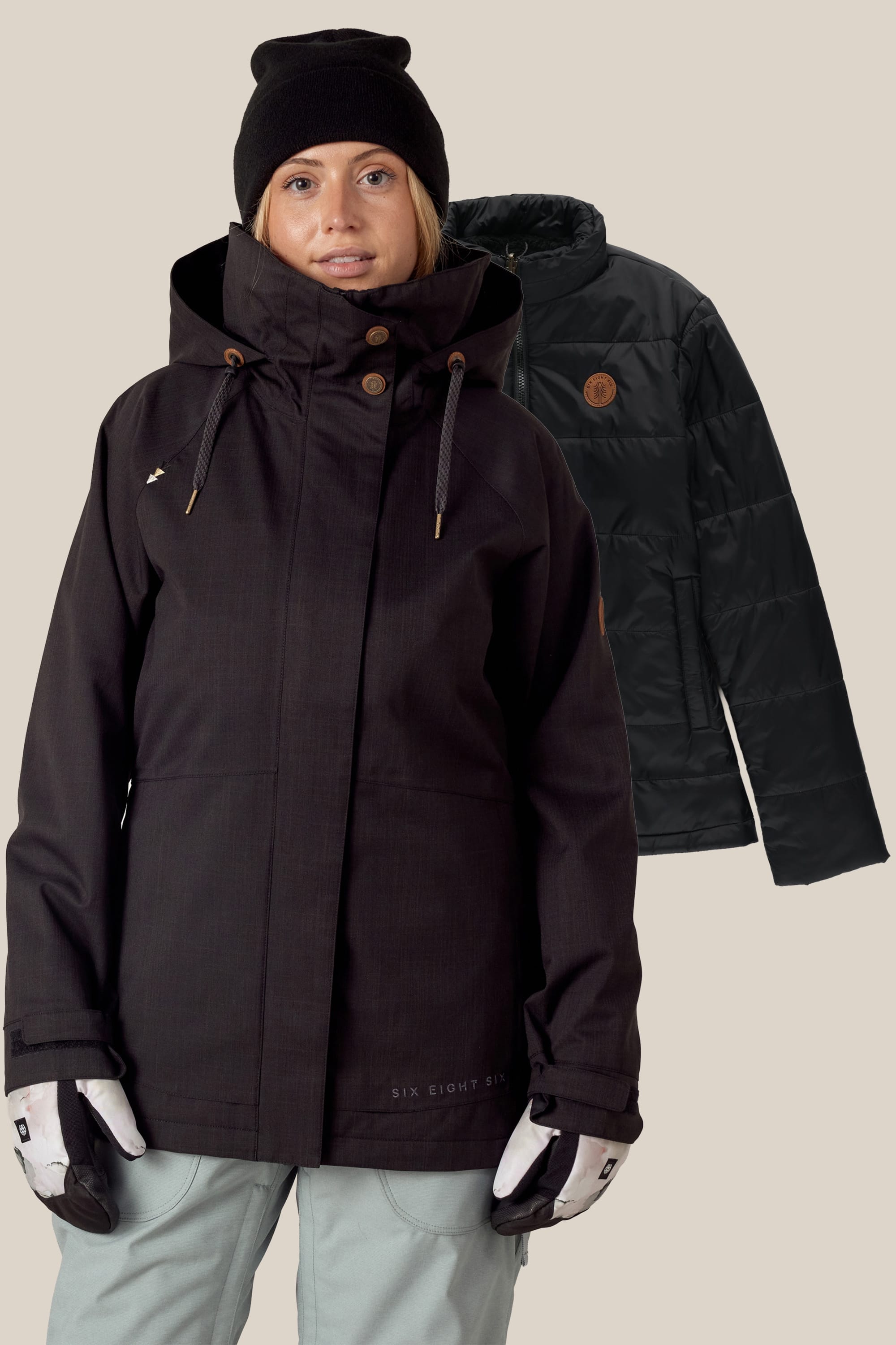 A woman in a black winter jacket, light gray pants, white gloves, and a black beanie stands before a plain background. Behind her hangs the 686 Women's SMARTY® 3-In-1 Spellbound Jacket by 686.