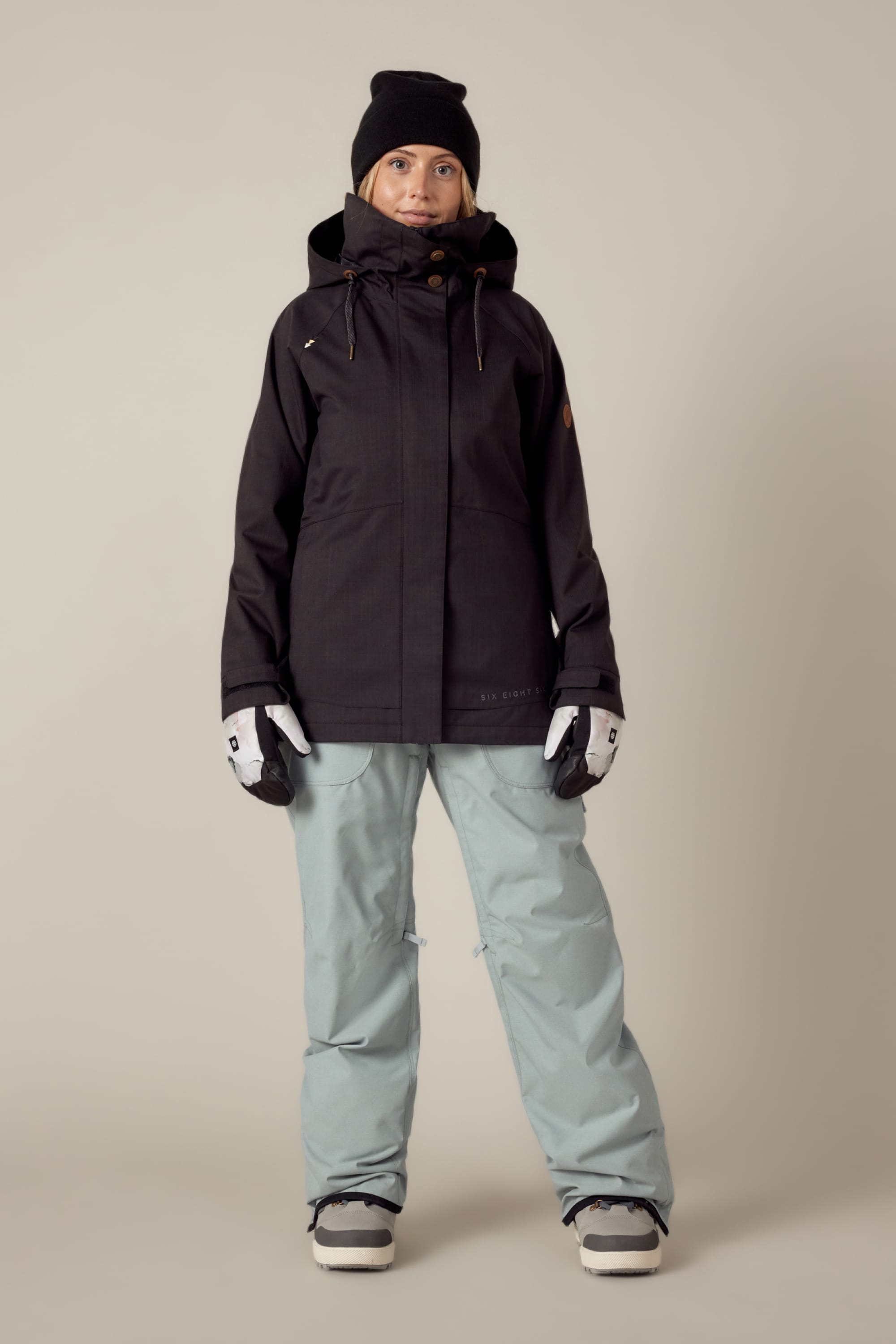 A person stands indoors against a plain beige background, wearing the 686 Women's SMARTY® 3-In-1 Spellbound Jacket in black, light blue snow pants, a black beanie, white gloves, and snow boots, looking directly at the camera.