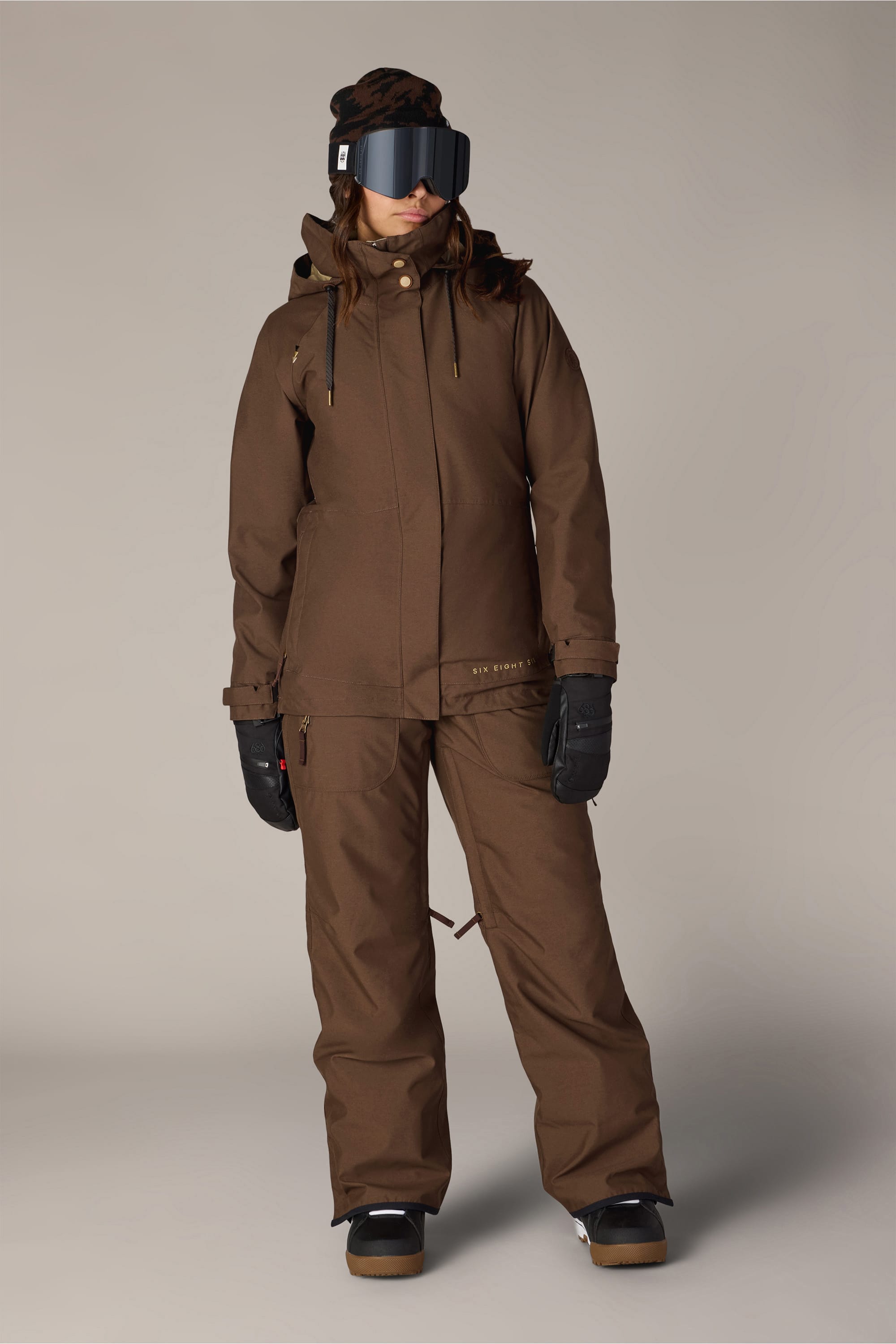 A person stands against a plain background wearing the 686 Women’s SMARTY® 3-In-1 Spellbound Jacket, along with matching pants, black gloves, dark boots, and large black ski goggles.