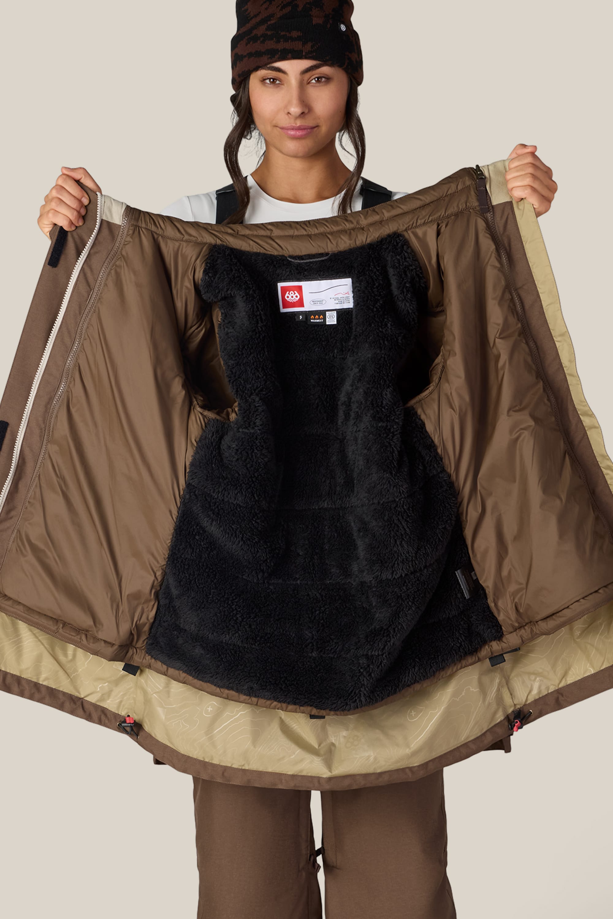 A person wearing brown pants and a beanie opens the 686 Women’s SMARTY® 3-In-1 Spellbound Jacket by 686 to reveal its black furry lining and interior details against a plain background.