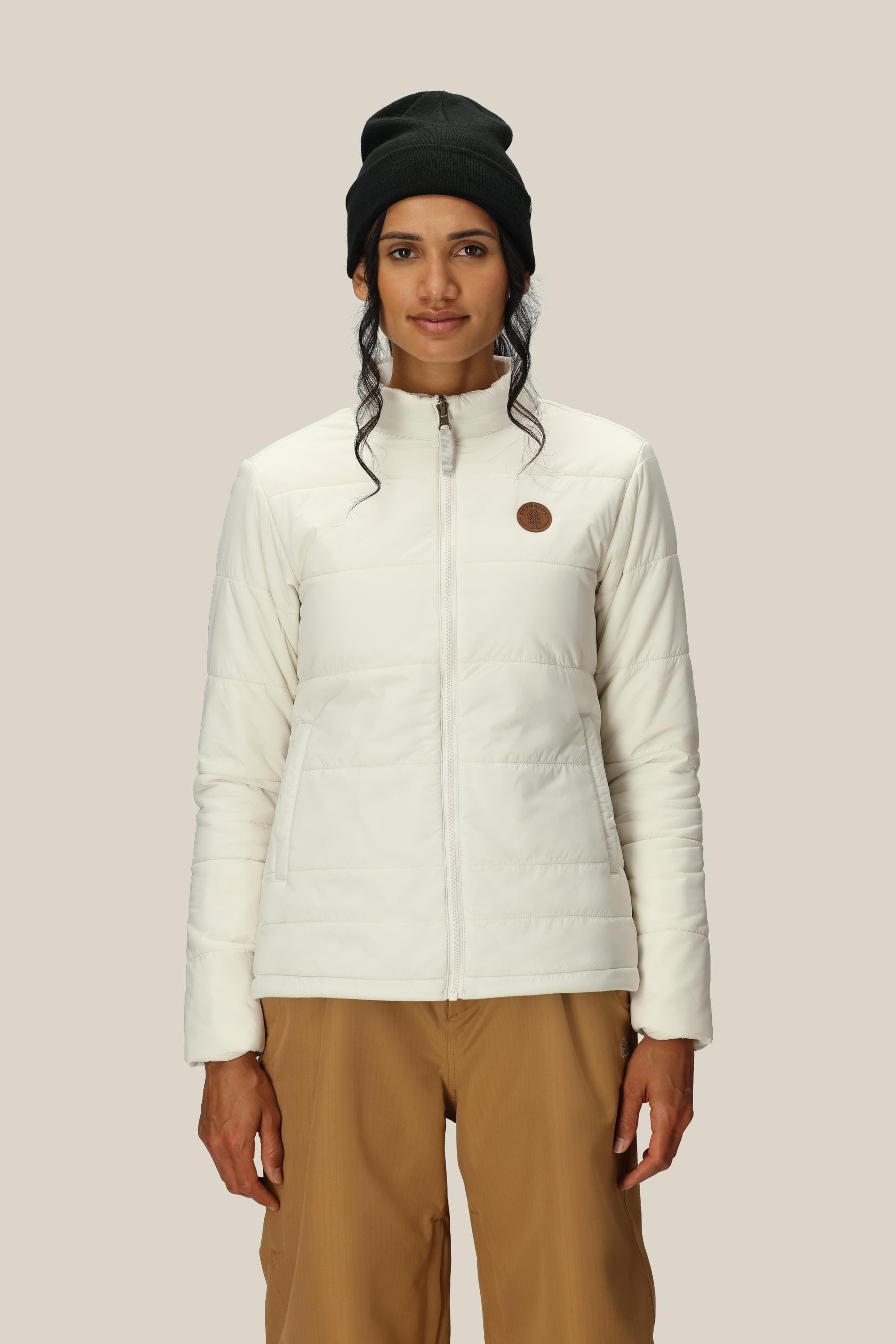 A person stands facing forward against a plain beige background, wearing the 686 Women's SMARTY® 3-In-1 Spellbound Jacket by 686 in white, paired with a black beanie and light brown pants.
