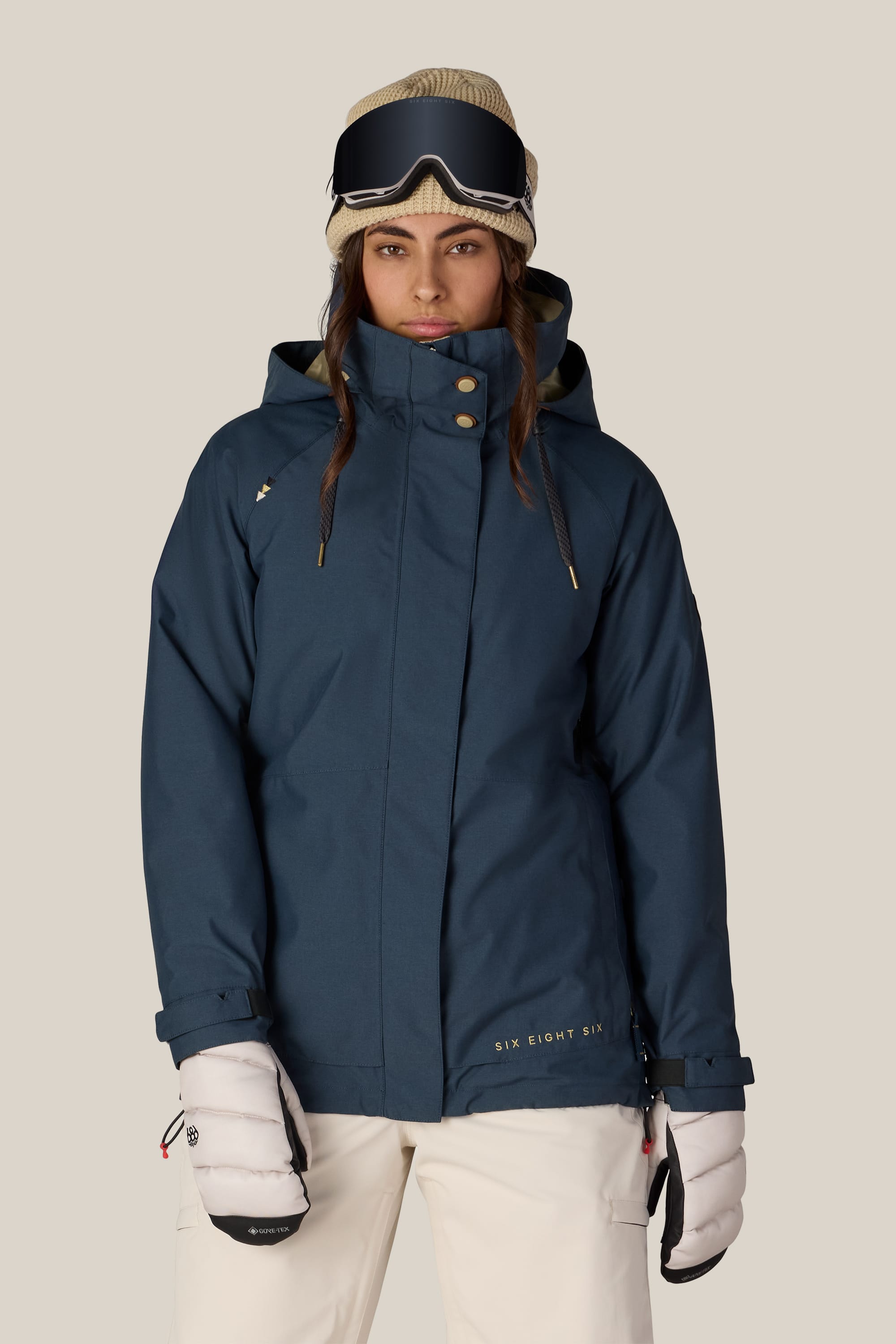 A woman wearing the 686 Women’s SMARTY® 3-In-1 Spellbound Jacket by 686, paired with white gloves, a beige hat, and ski goggles, stands facing forward against a plain background.