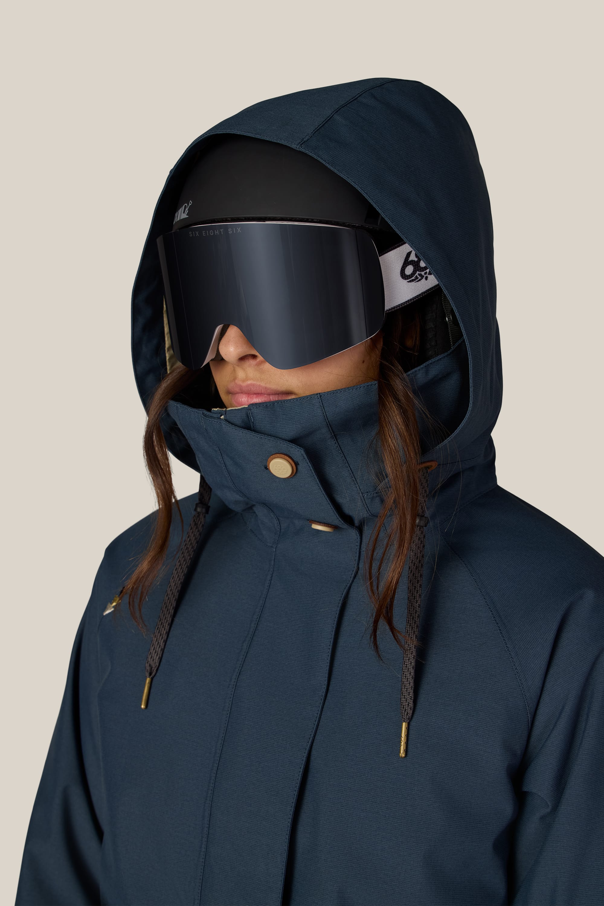 A woman wearing the 686 Women’s SMARTY® 3-In-1 Spellbound Jacket by 686, along with a black ski helmet and large reflective ski goggles, her brown hair peeking out, stands against a neutral background.