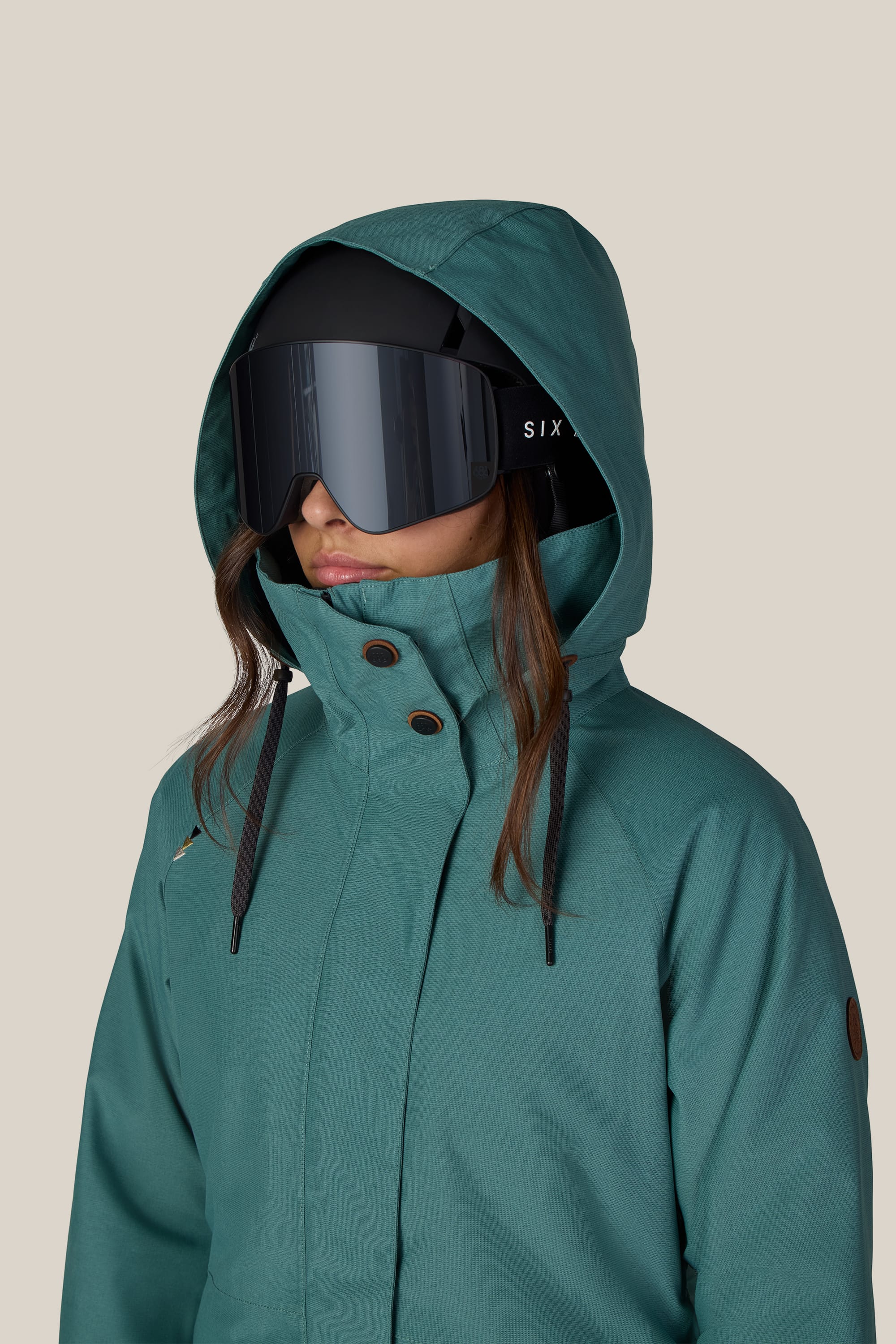 A person wearing the 686 Women’s SMARTY® 3-In-1 Spellbound Jacket and large black ski goggles stands against a plain beige background, facing slightly to the side.