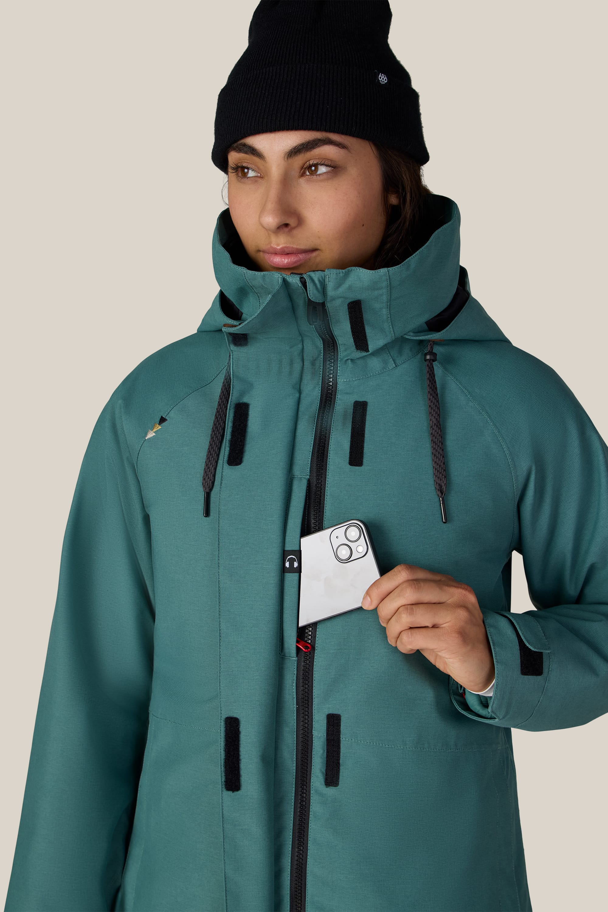 A woman wearing the 686 Women’s SMARTY® 3-In-1 Spellbound Jacket in teal and a black beanie places her smartphone into a chest pocket, standing against a plain light background.