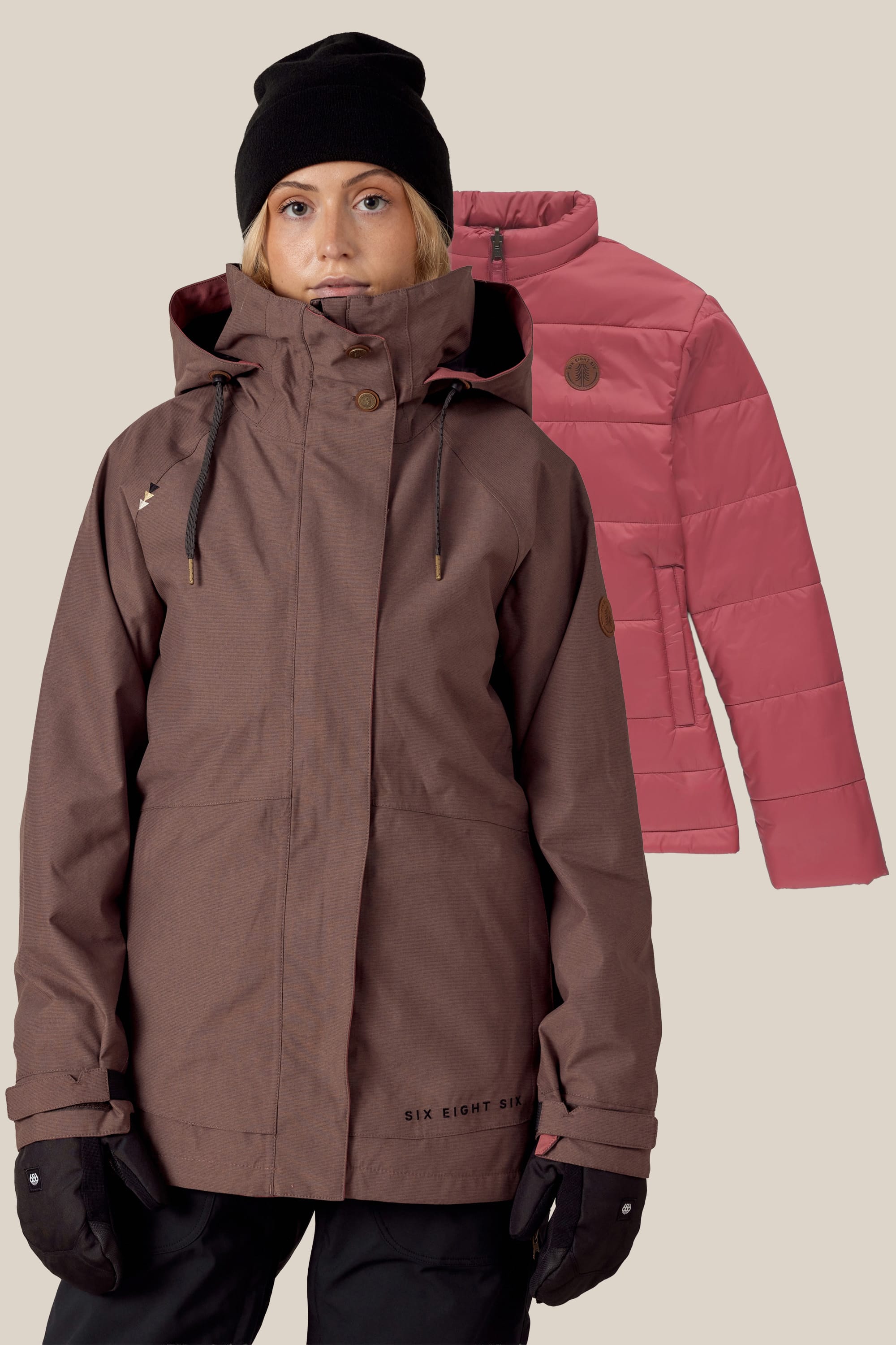 A person in a brown winter jacket, black beanie, and gloves stands before the 686 Women's SMARTY® 3-In-1 Spellbound Jacket by 686, featured in pink against a light beige backdrop.