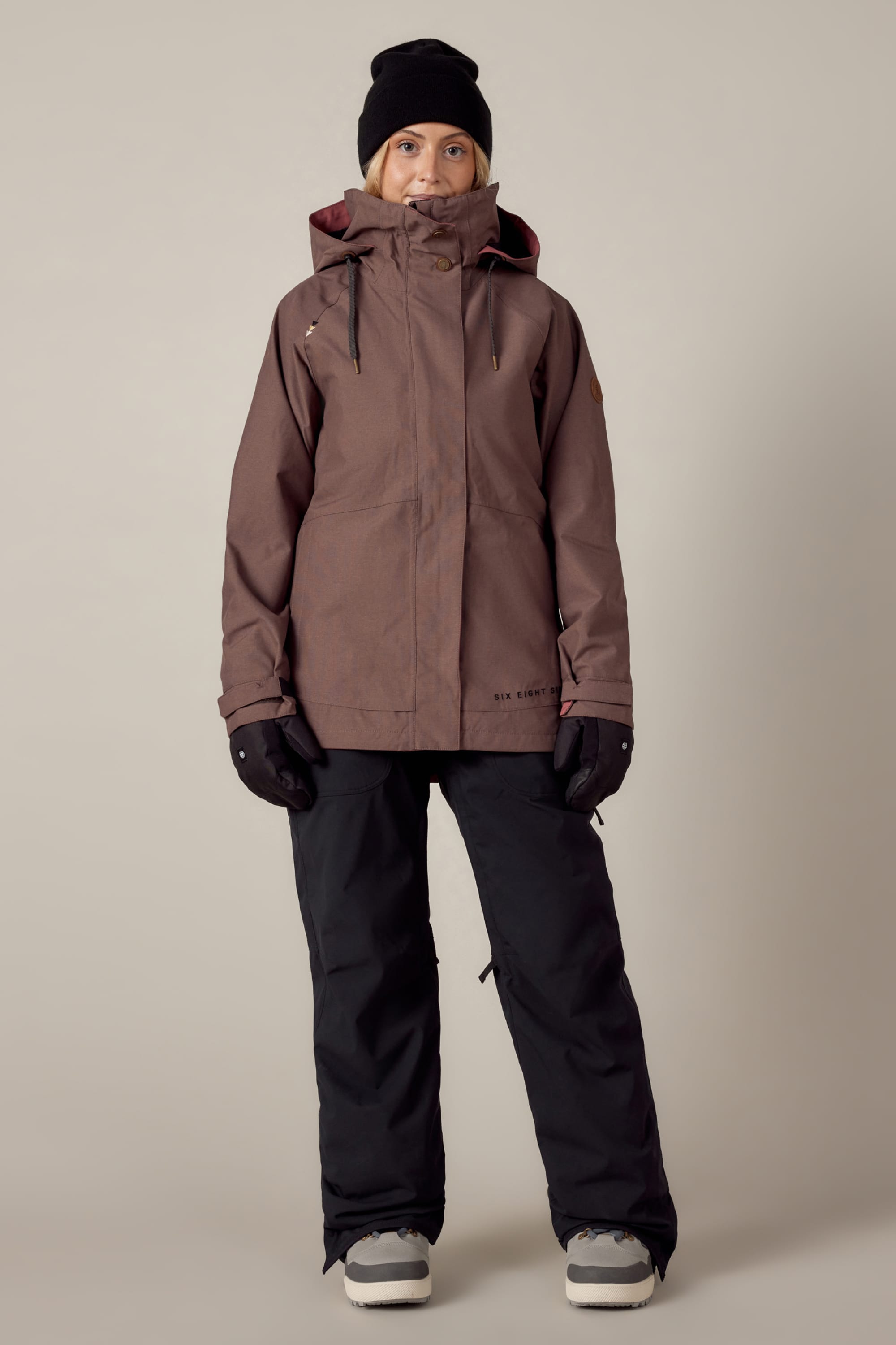 A person stands facing forward wearing the 686 Women's SMARTY® 3-In-1 Spellbound Jacket with a hood by 686, paired with a black beanie, black gloves, black pants, and gray winter boots against a plain light background.