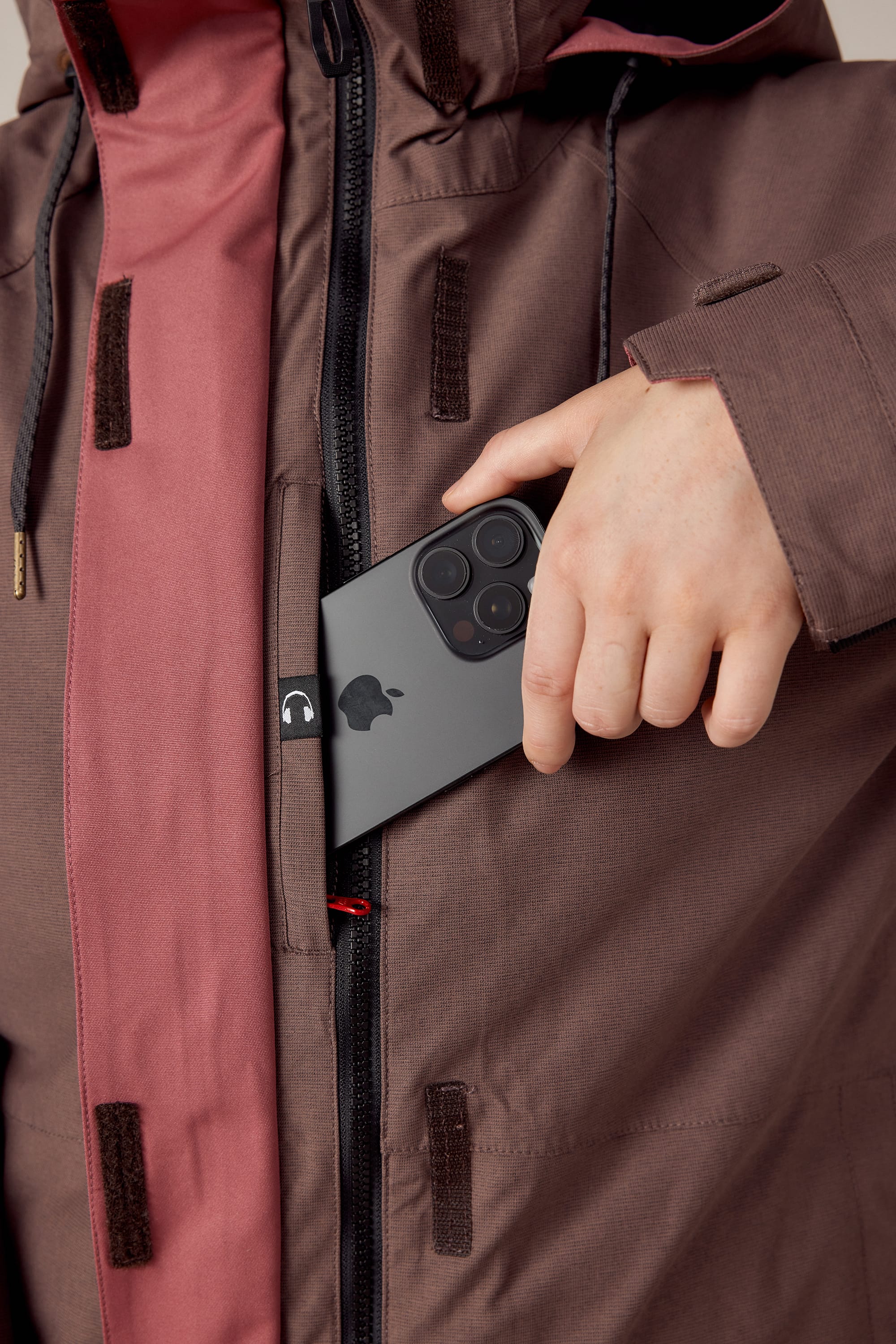 A woman wearing the 686 Women's SMARTY® 3-In-1 Spellbound Jacket places a black iPhone with three camera lenses into an inner chest pocket of the waterproof brown and pink jacket.