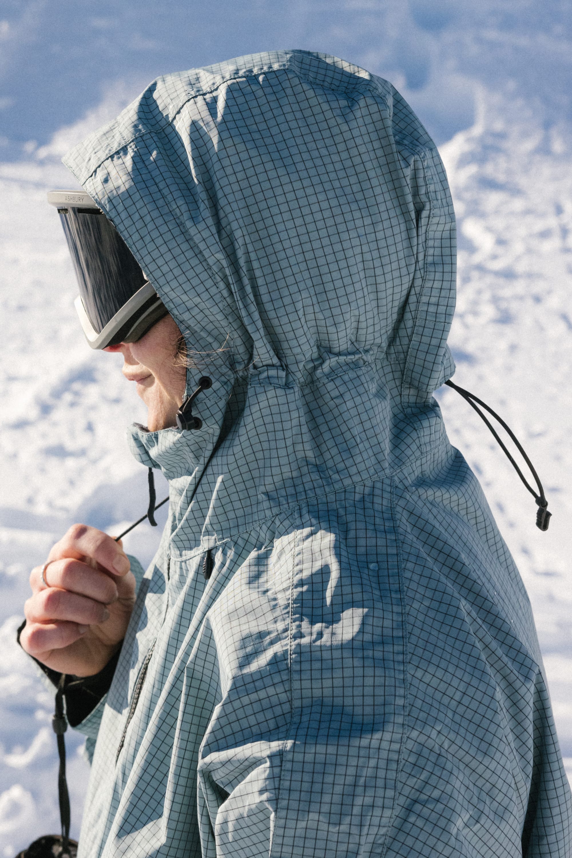 A person wearing the 686 Women's Outline™ Shell Anorak and ski goggles stands in a snowy outdoor setting, pulling the hood drawstring.