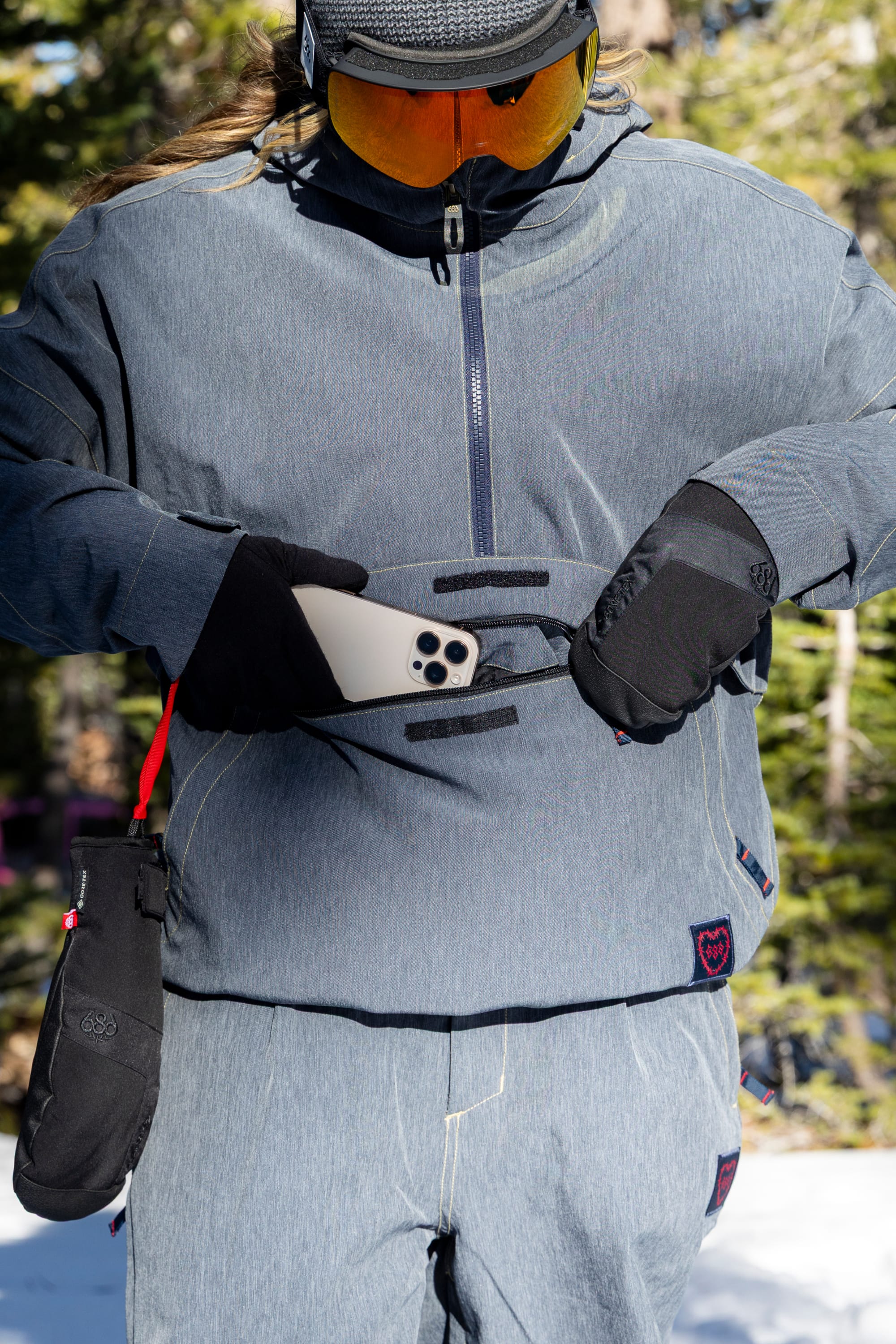 Wearing the 686 Women’s Outline™ Shell Anorak by 686, a person in gray winter gear and orange goggles slips a smartphone into the jacket’s zippered pocket, while a black pouch hangs from their wrist. Snow and trees fill the background.