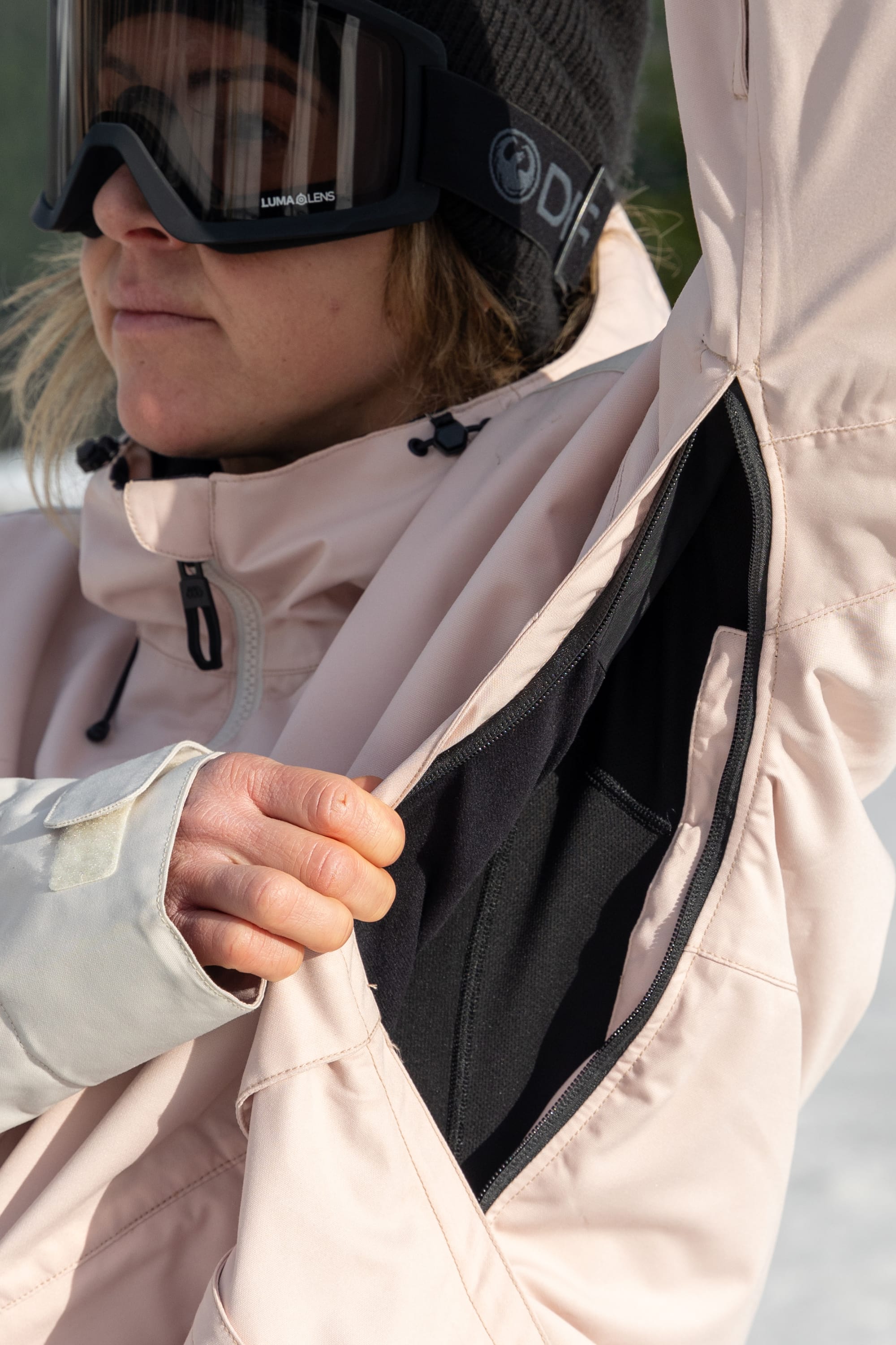 A person in the 686 Women's Outline™ Shell Anorak, a light pink waterproof jacket, unzips the underarm vent to show a black inner layer. Ski goggles and a snowy, tree-filled background complete the look.