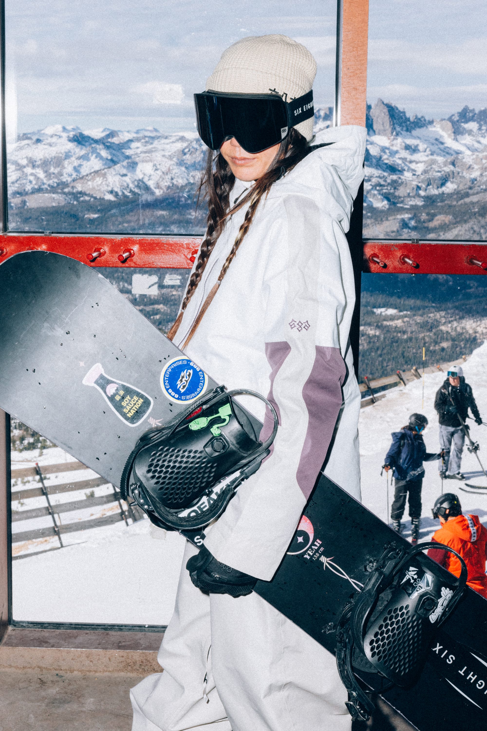 Wearing the 686 Women’s Outline™ Shell Anorak from 686 and goggles, a woman holds a snowboard indoors while a snowy mountain landscape and people dressed in winter clothing are visible through the window in the background.