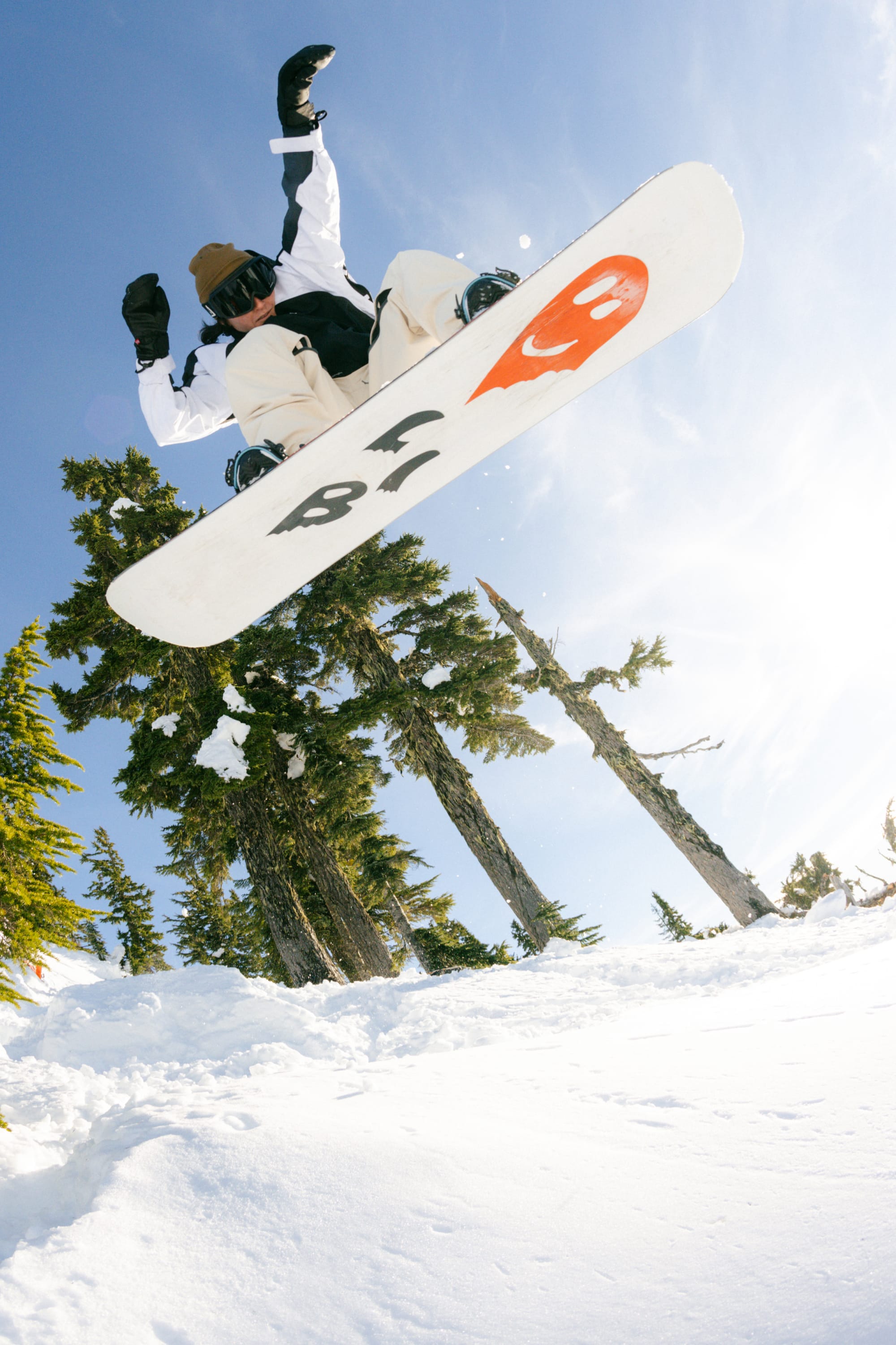 A snowboarder in a brown beanie and a 686 Women's Hologram™ Shell Jacket by 686 catches air mid-jump on a snowy mountain, surrounded by tall evergreens beneath a bright blue sky.