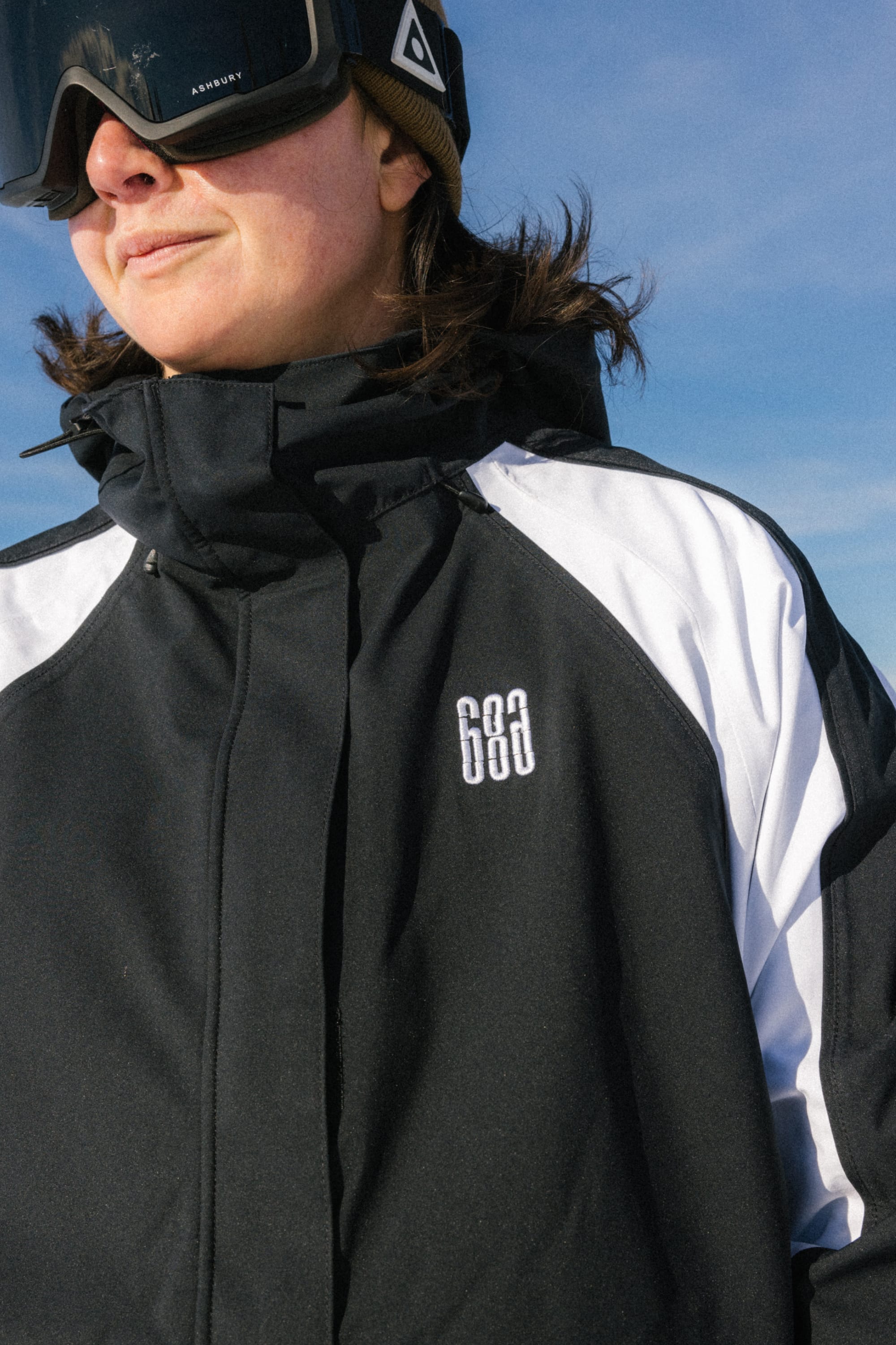 A person in the 686 Women's Hologram™ Shell Jacket and black ski goggles wears a brown hat outdoors under a blue sky.