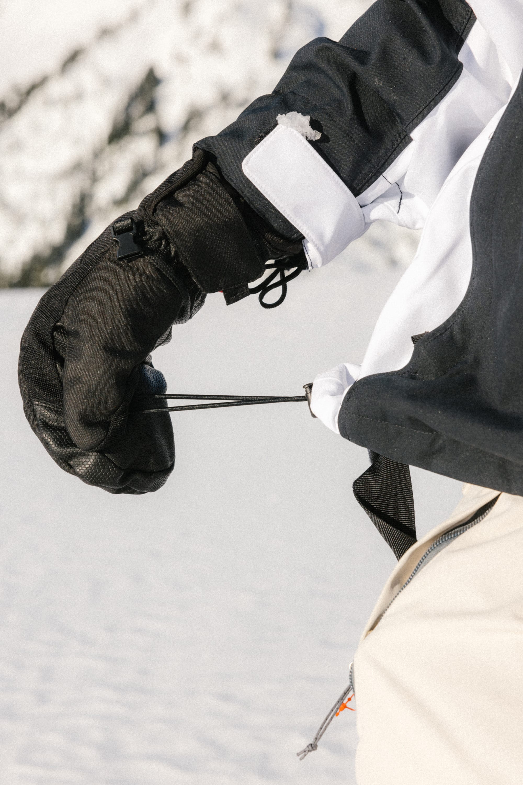 A person wearing black gloves and a 686 Women's Hologram™ Shell Jacket adjusts the drawstring on their coat, with snowy mountains in the background.