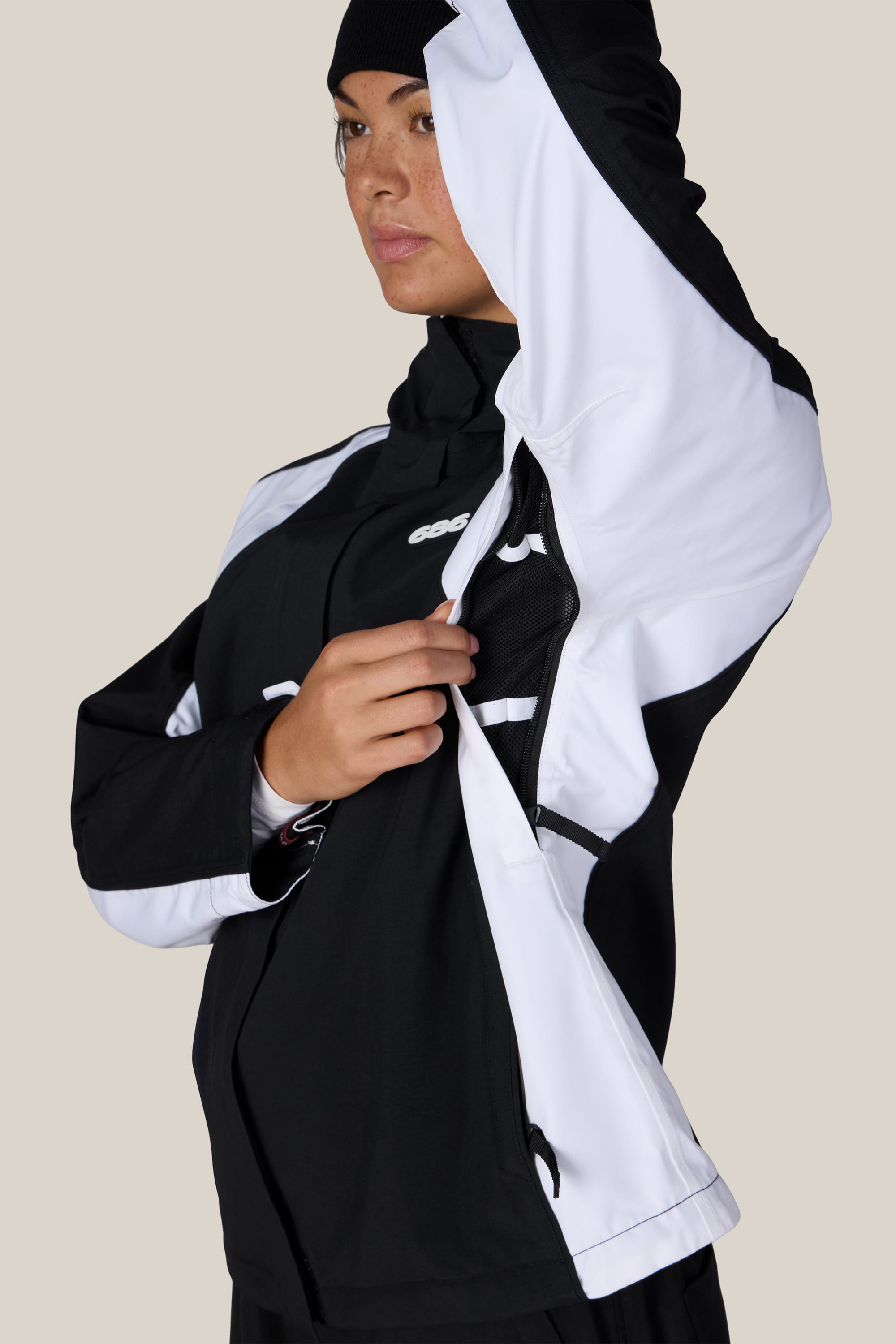 A woman wearing the 686 Women’s Hologram™ Shell Jacket in black and white unzips a ventilation opening beneath the arm, revealing mesh lining inside. She is also sporting a black beanie and looking slightly to the side.