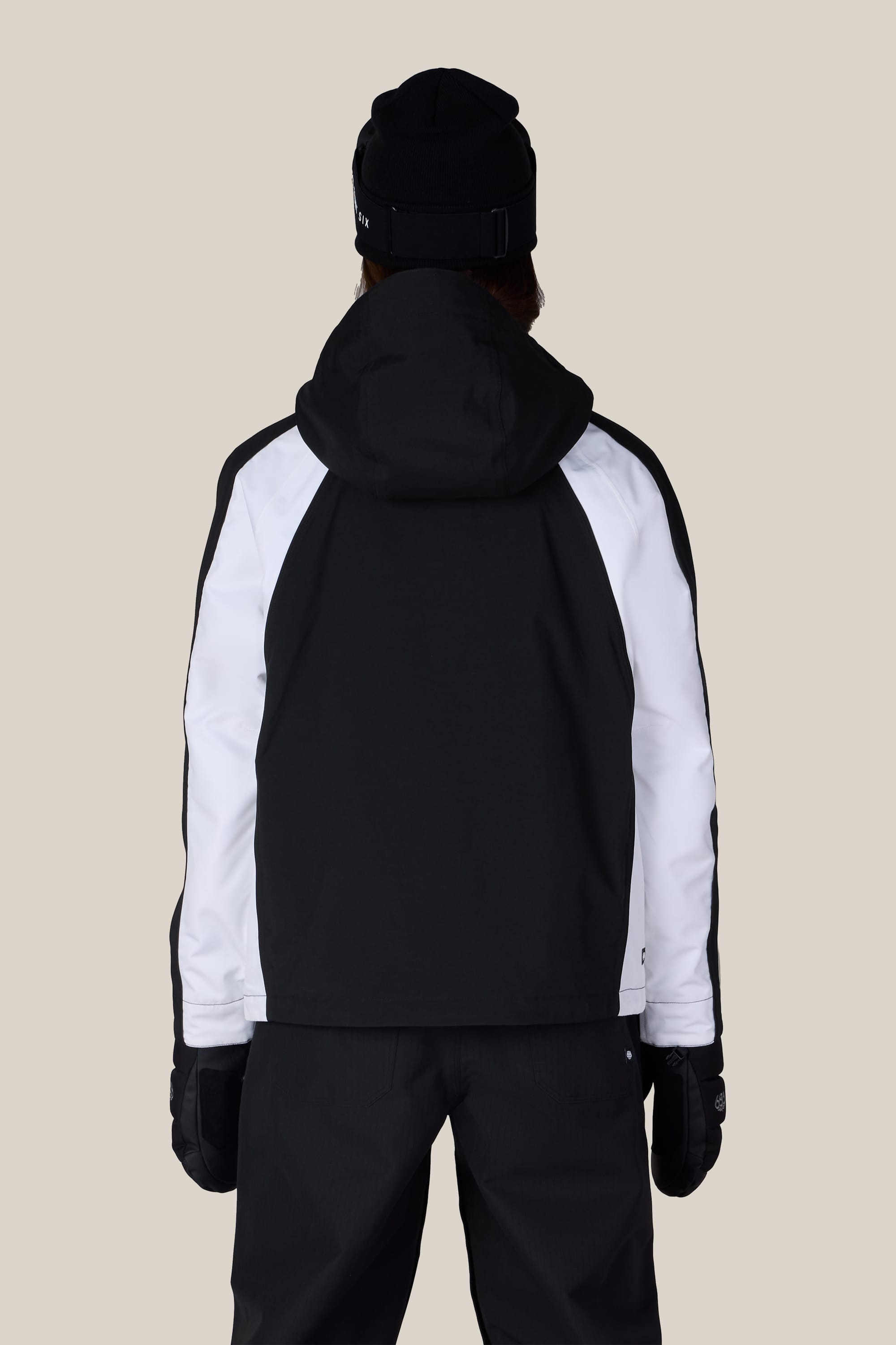 A person wearing the 686 Women’s Hologram™ Shell Jacket by 686, along with black pants, a black beanie, and black gloves, stands facing away from the camera against a plain beige background.