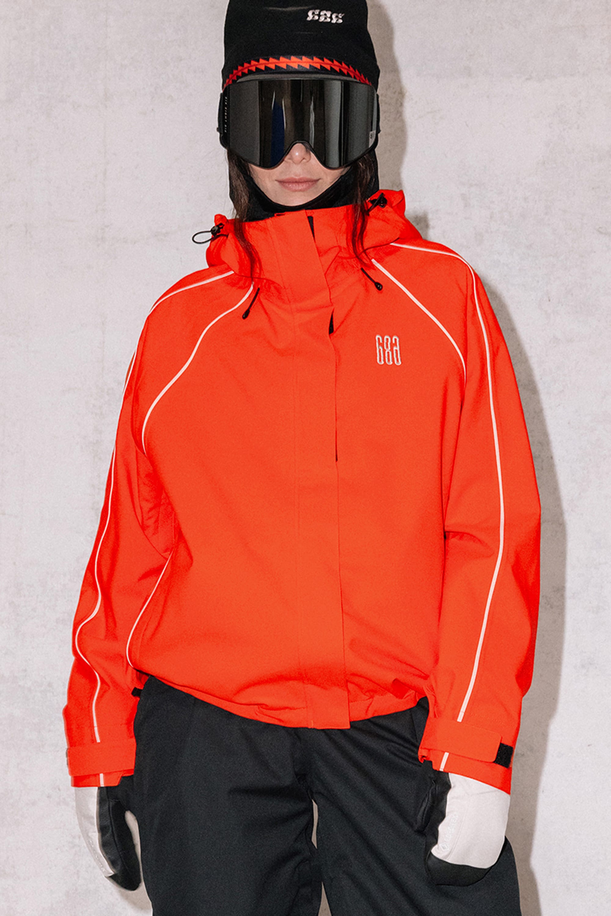 A person in a 686 Women's Hologram™ Shell Jacket, black pants, white gloves, black beanie, and black ski goggles stands against a plain light-colored wall.