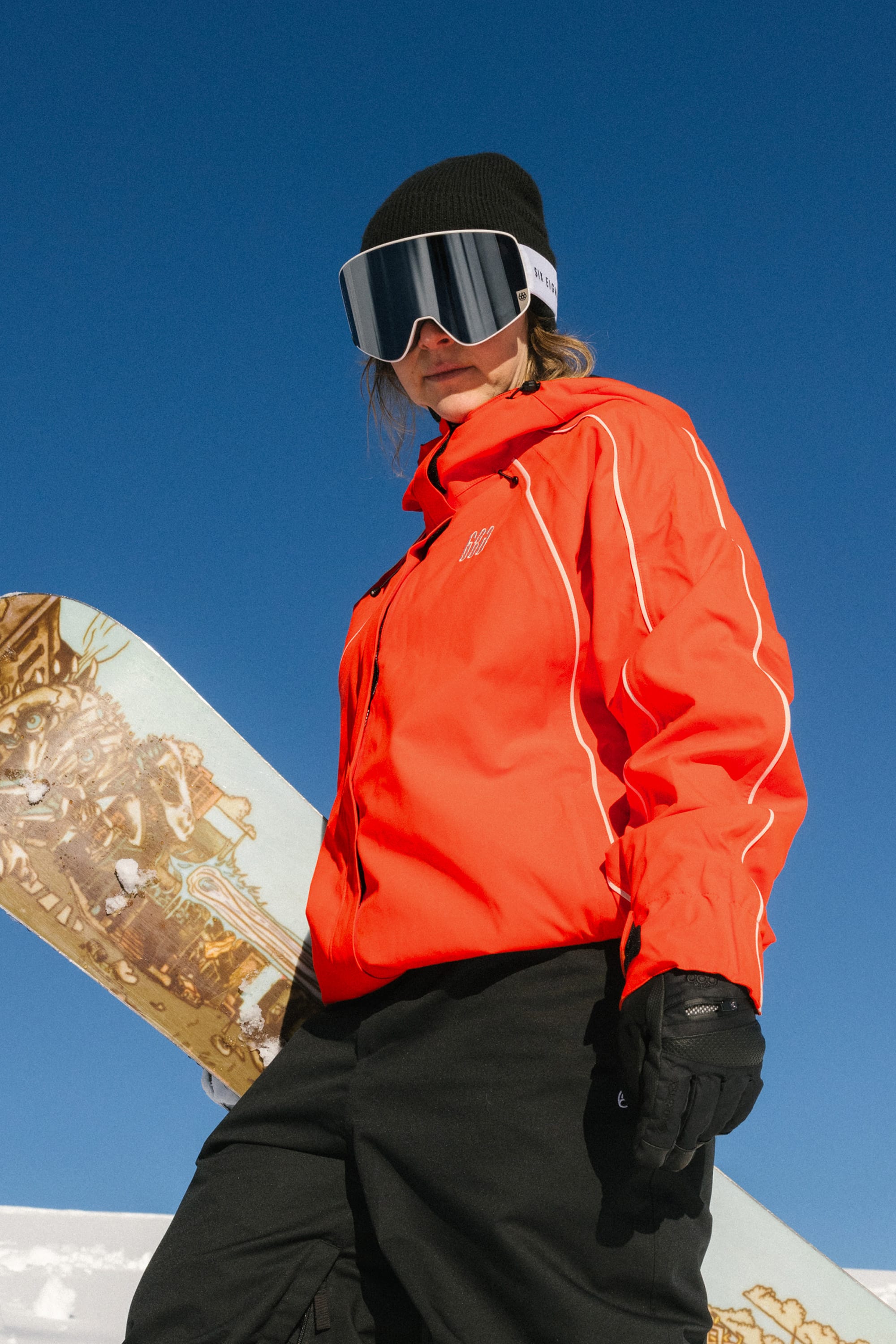 Wearing the 686 Women's Hologram™ Shell Jacket and a black beanie, a woman in ski goggles and gloves stands outdoors holding a snowboard against a clear blue sky, looking confidently at the camera.