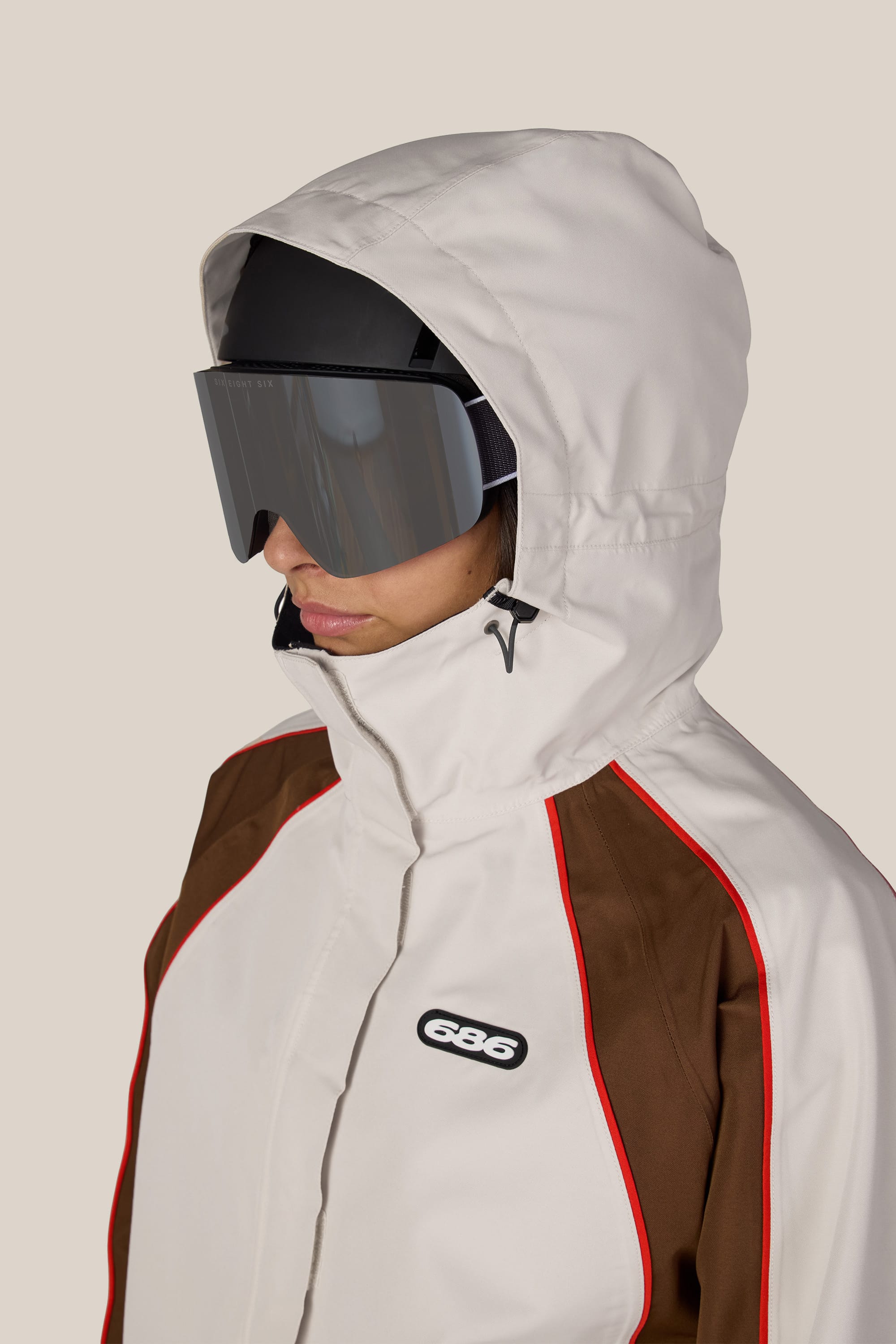 A person wearing the 686 Women’s Hologram™ Shell Jacket—a white hooded jacket with brown panels and red piping—from 686, paired with large black ski goggles and a black helmet, is facing left against a plain background.