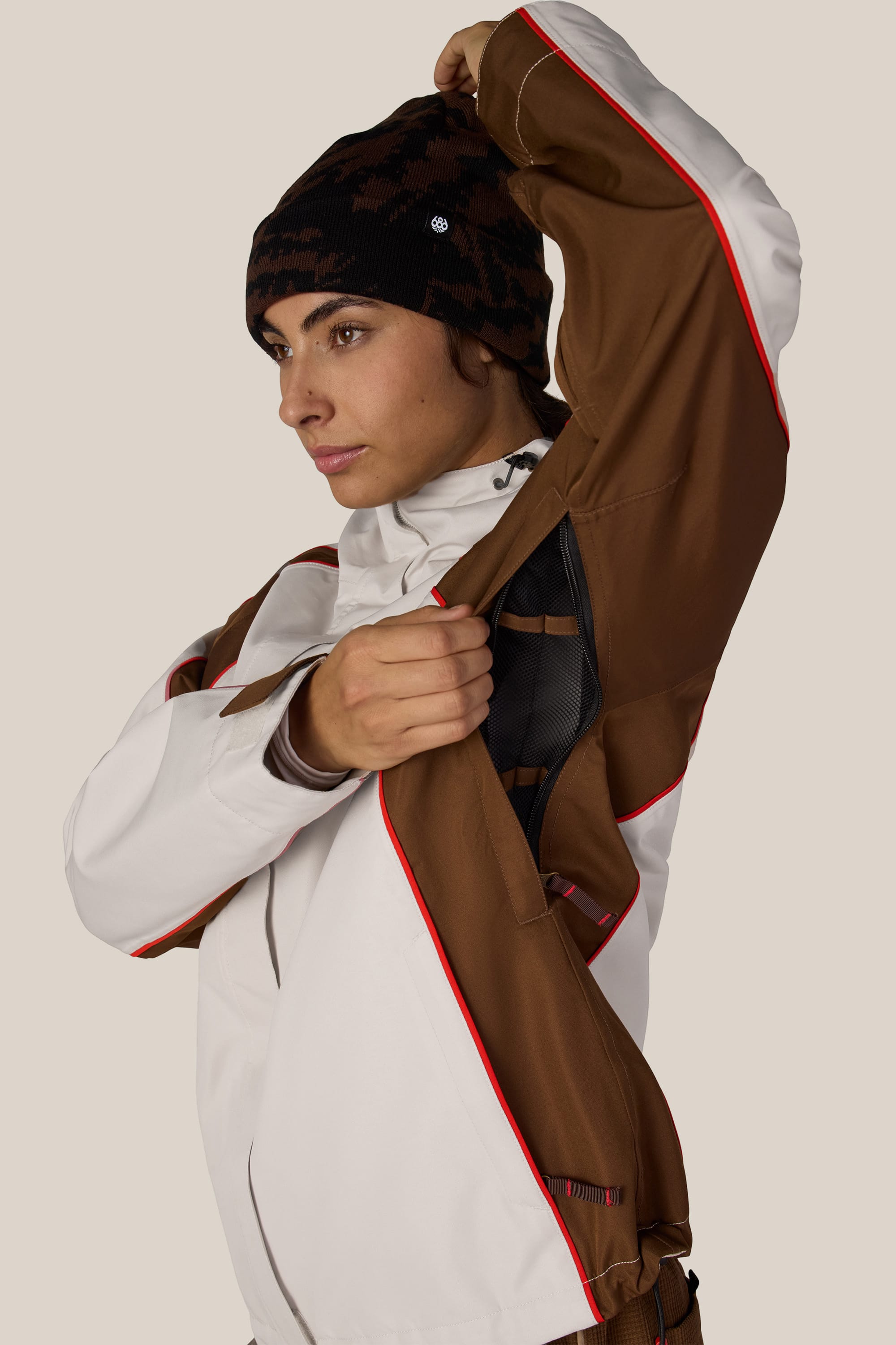 A person wearing the 686 Women’s Hologram™ Shell Jacket in white and brown with red accents unzips a side vent while sporting a black patterned beanie.