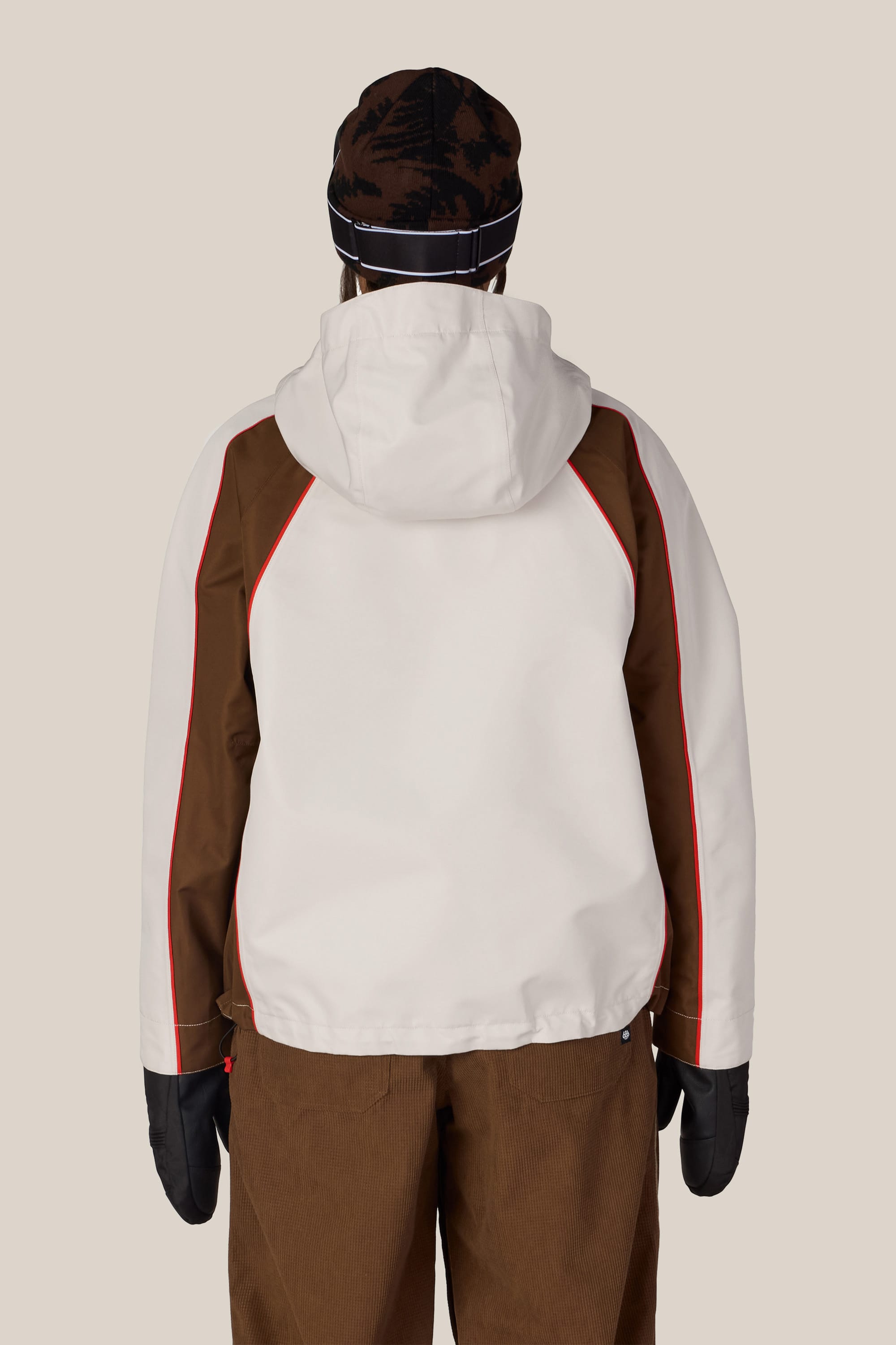 A person facing away, wearing the 686 Women’s Hologram™ Shell Jacket in white and brown with red accents, brown pants, black mittens, and a black and brown beanie. The background is a plain, light beige color.