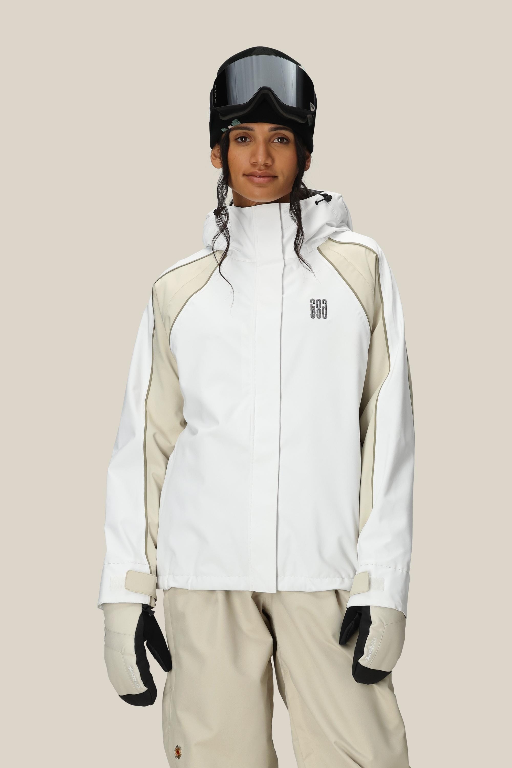 A woman wearing the 686 Women's Hologram™ Shell Jacket in white and beige stands facing forward against a plain light background, paired with beige ski pants, white gloves, and black ski goggles.