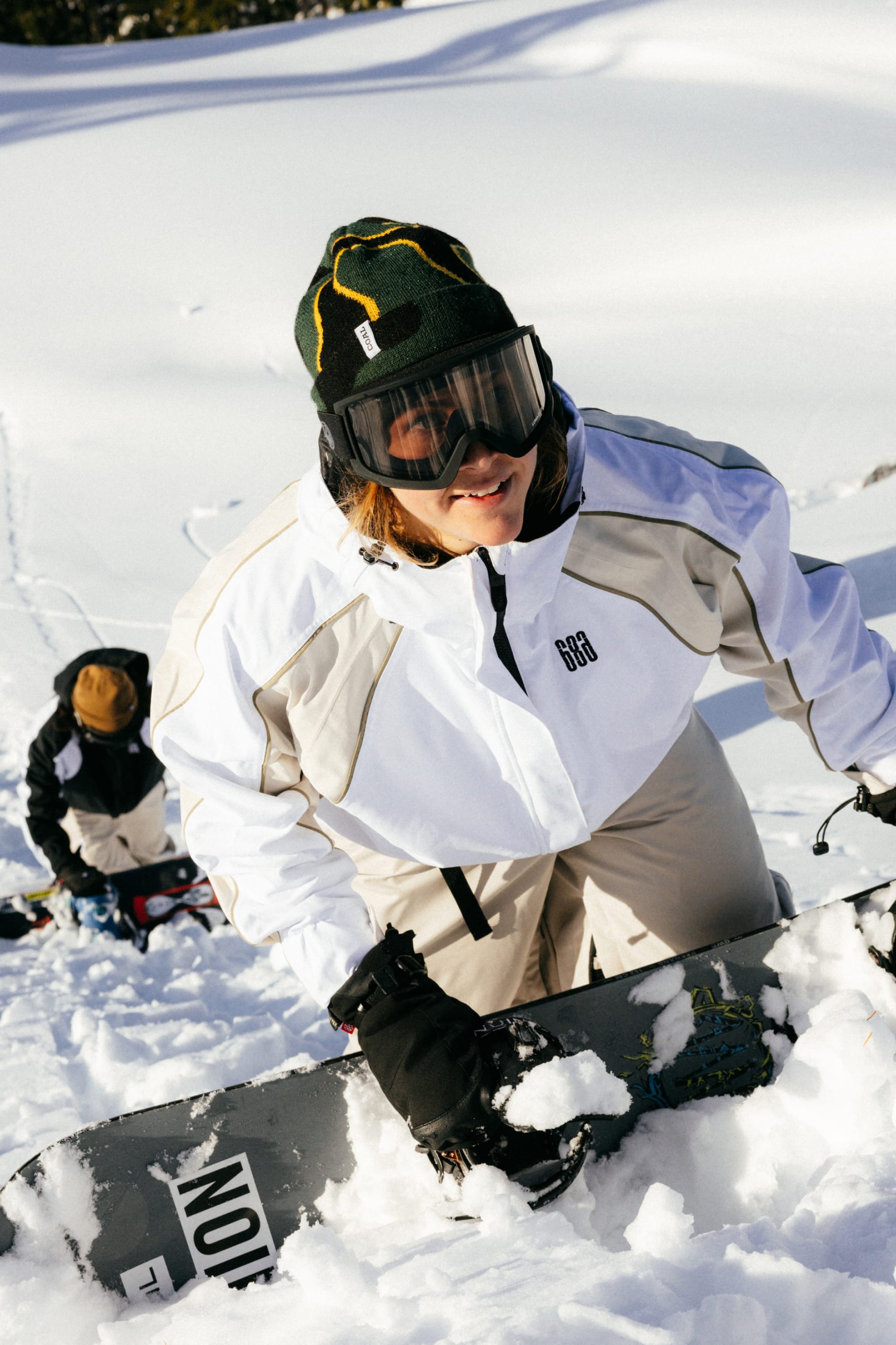 Two people in winter gear climb a snowy slope with snowboards. In front, someone wearing the 686 Women's Hologram™ Shell Jacket by 686, black gloves, goggles, and a green beanie smiles as they ascend through the snow-covered landscape.