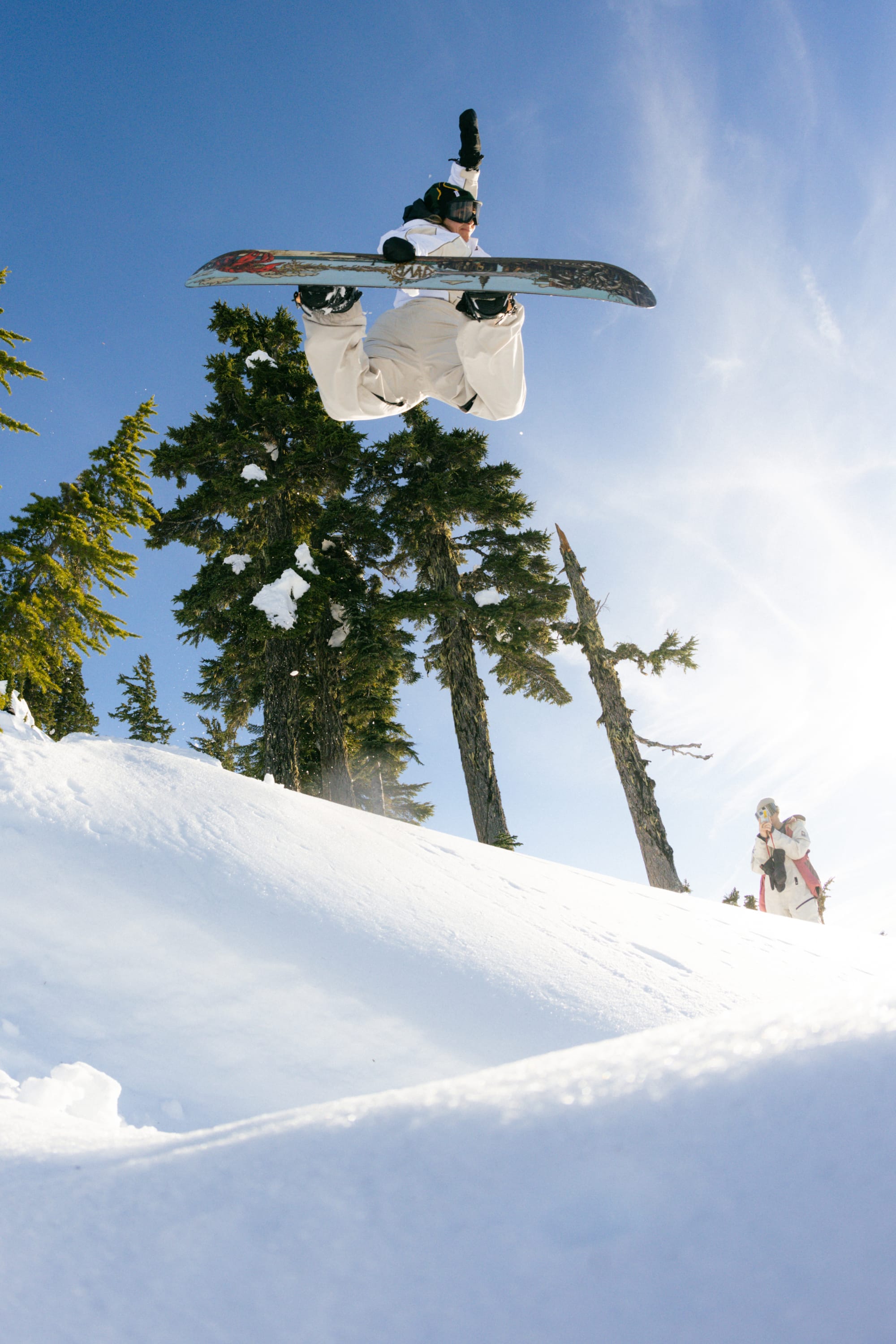 A snowboarder wearing the 686 Women's Hologram™ Shell Jacket by 686 soars mid-air above a snowy slope, with green pine trees, another rider, and a clear blue sky in the background.