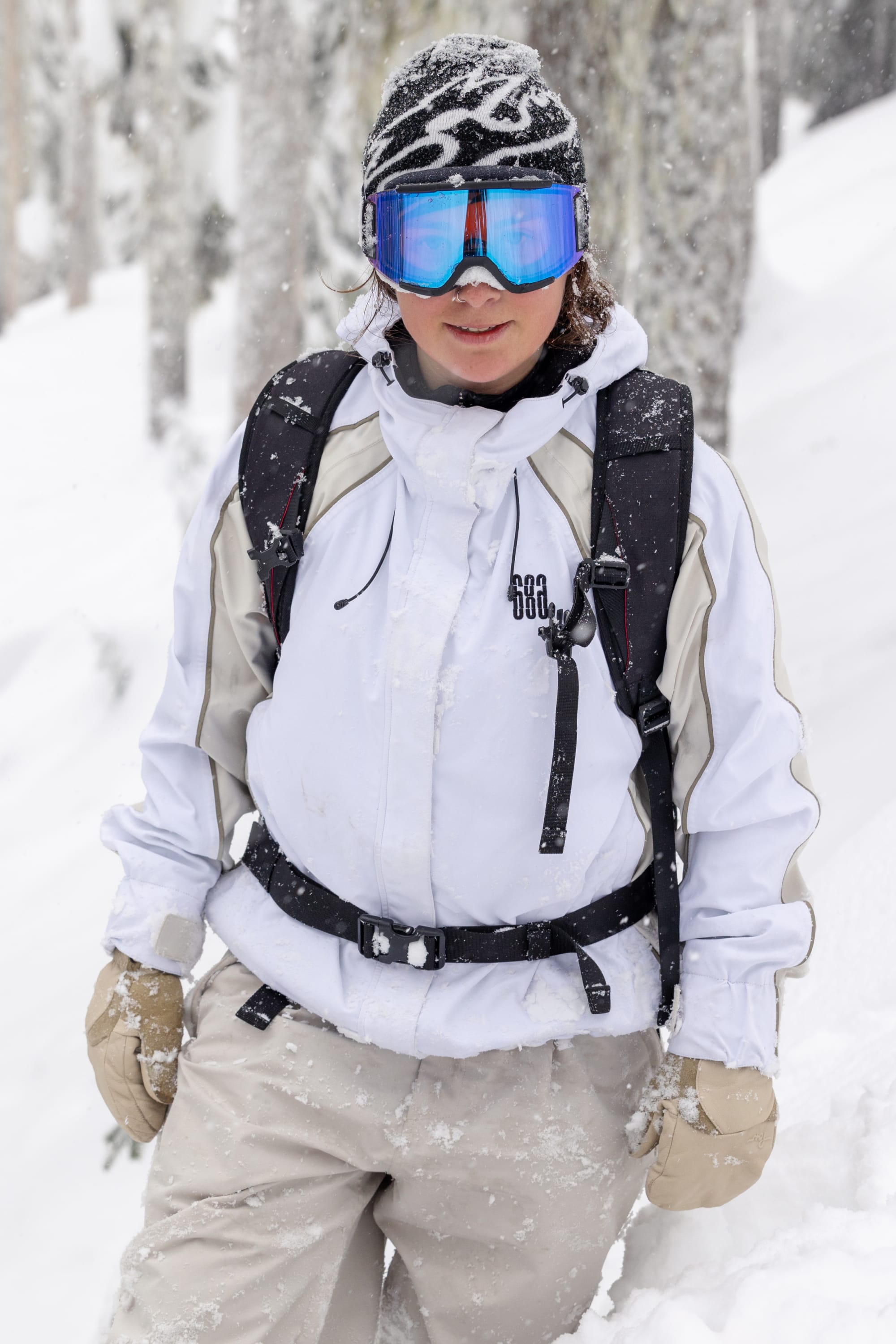 Wearing a 686 Women's Hologram™ Shell Jacket, beige pants, gloves, blue ski goggles, and a patterned beanie, a person stands in snowy woods with a black backpack as snow falls around them.