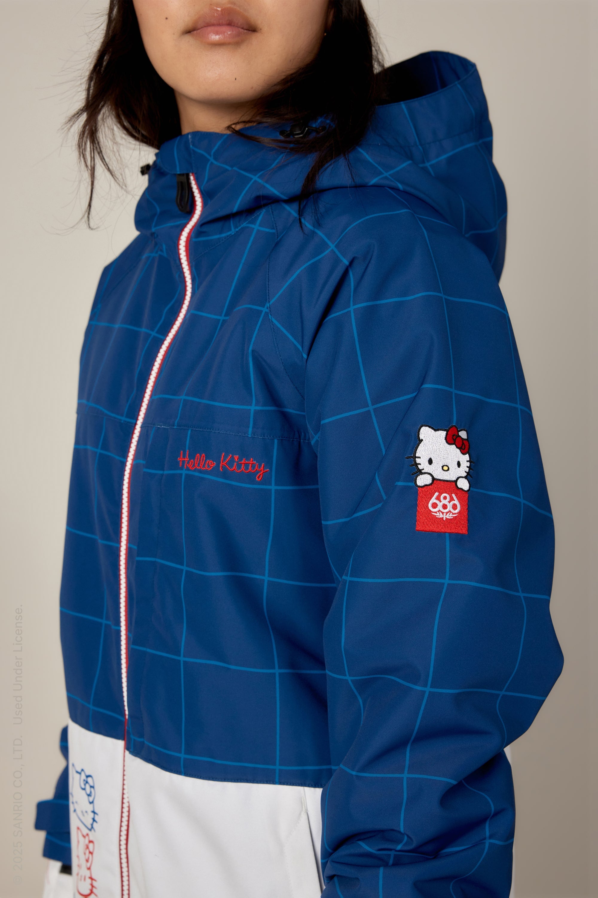 A person wears the 686 Women's Athena™ Insulated Jacket, a blue and white grid-patterned waterproof polyester piece by 686, featuring Hello Kitty embroidery on the chest, a sleeve patch, and a hood partly covering the face for playful charm.