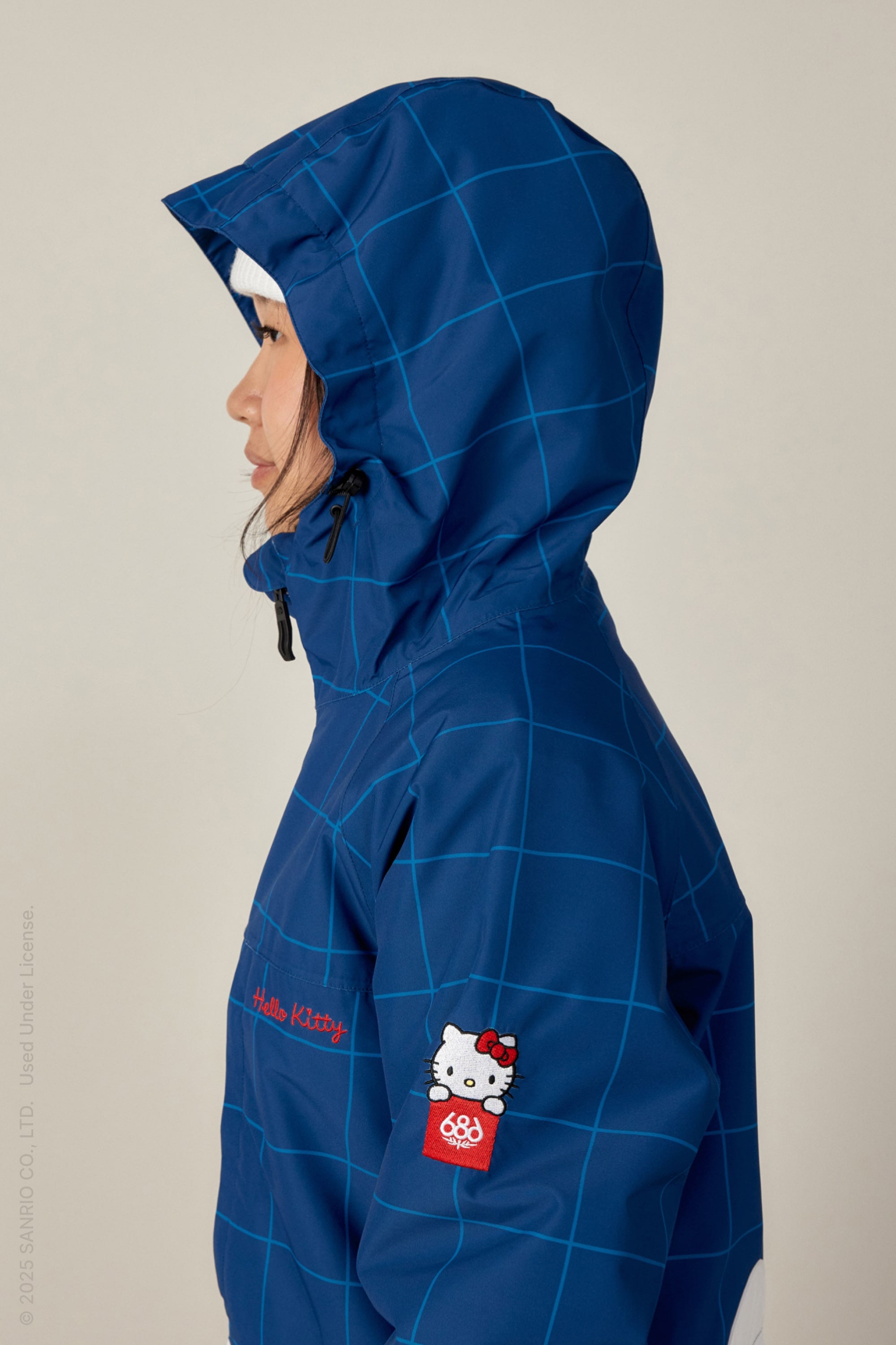 A person wears the 686 Women's Athena™ Insulated Jacket, a blue woven piece with a grid pattern and Hello Kitty patch on the sleeve, shown in profile against a plain background.