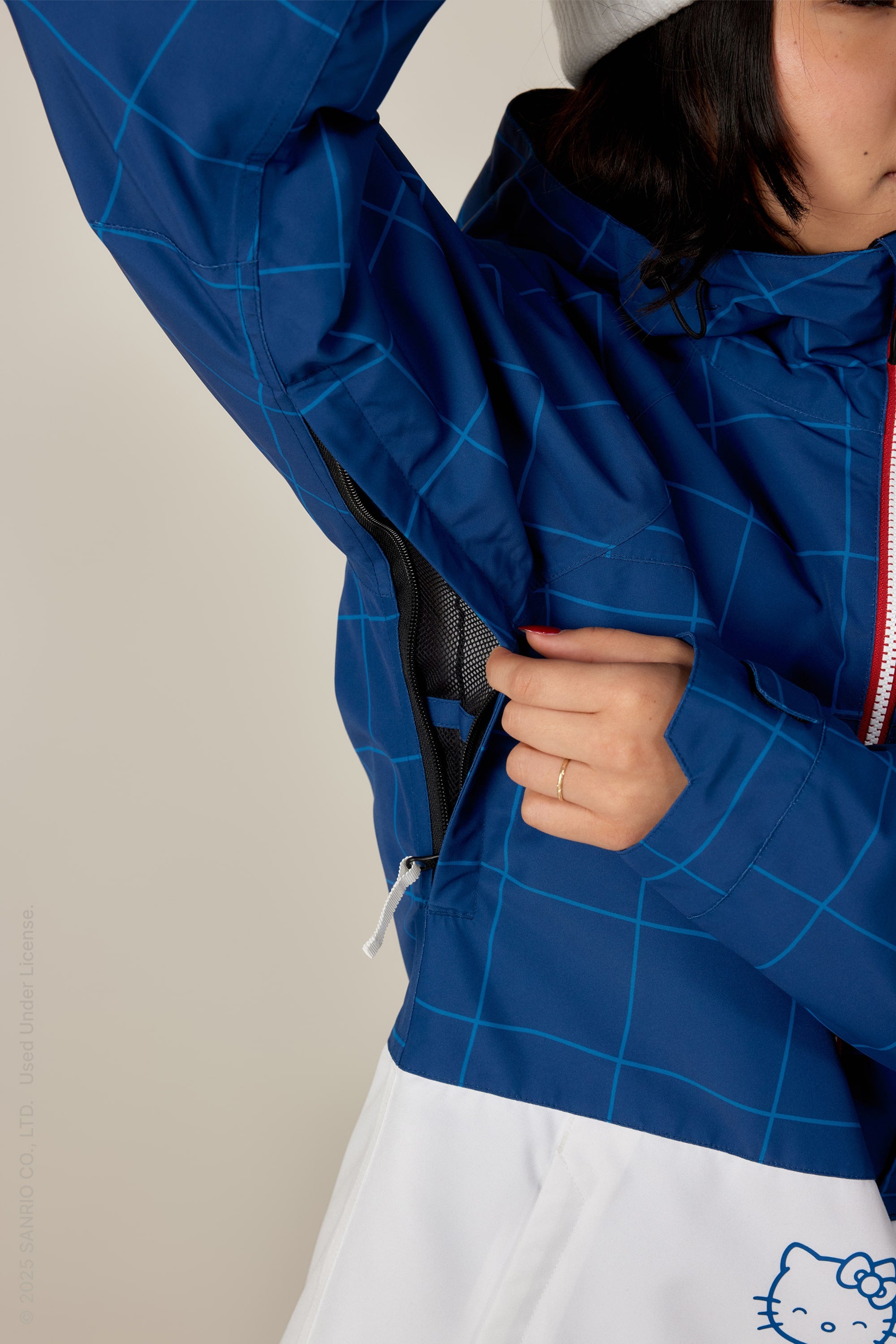 A person, partly visible, unzips a vent under the arm of a blue and white 686 Women's Athena™ Insulated Jacket featuring grid lines and a small Hello Kitty graphic near the hem. A gold ring is visible on their hand.