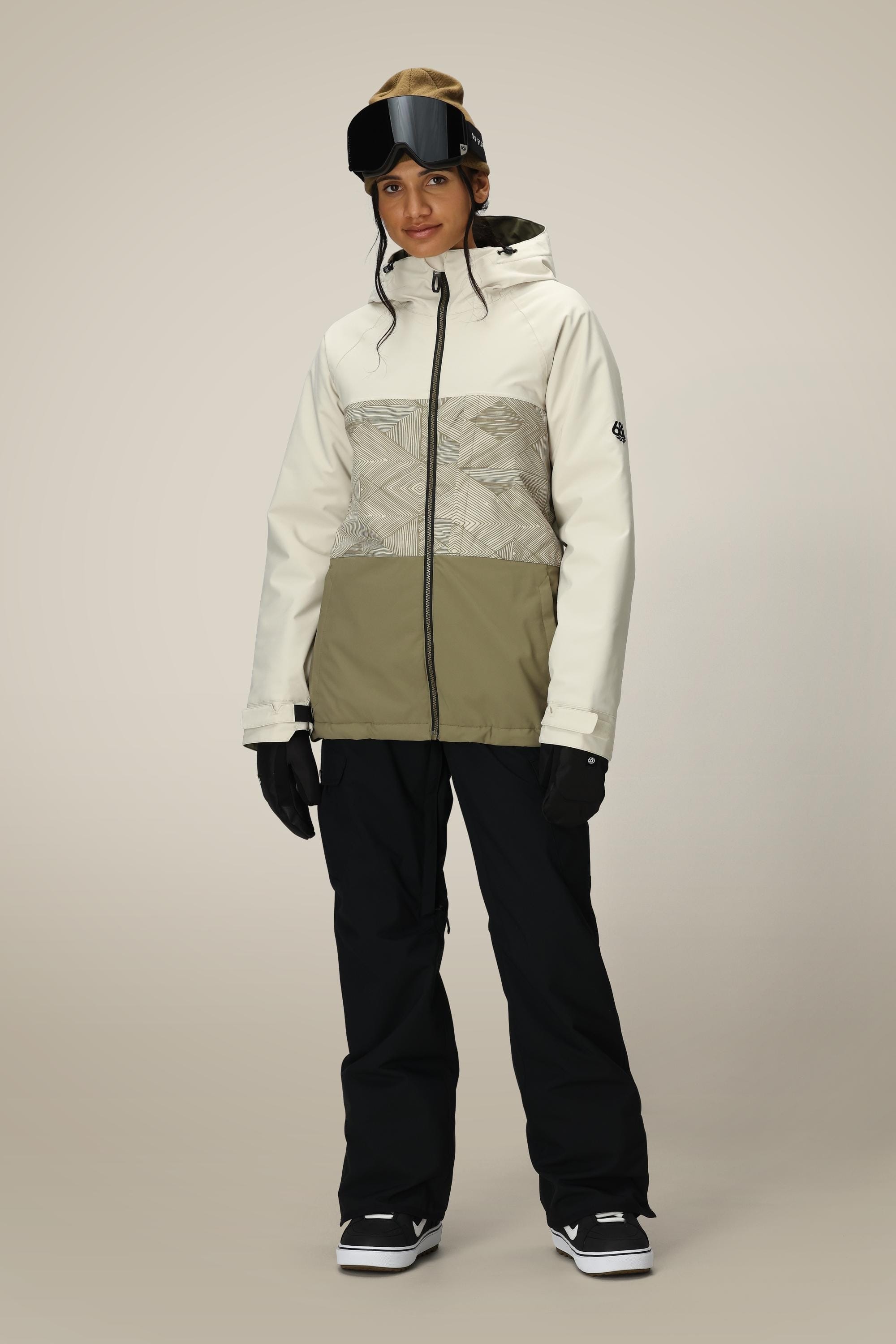 A person wears the 686 Women's Athena™ Insulated Jacket in beige and olive green, paired with black snow pants, gloves, black snow goggles on their head, and black snow boots, standing against a plain light background.