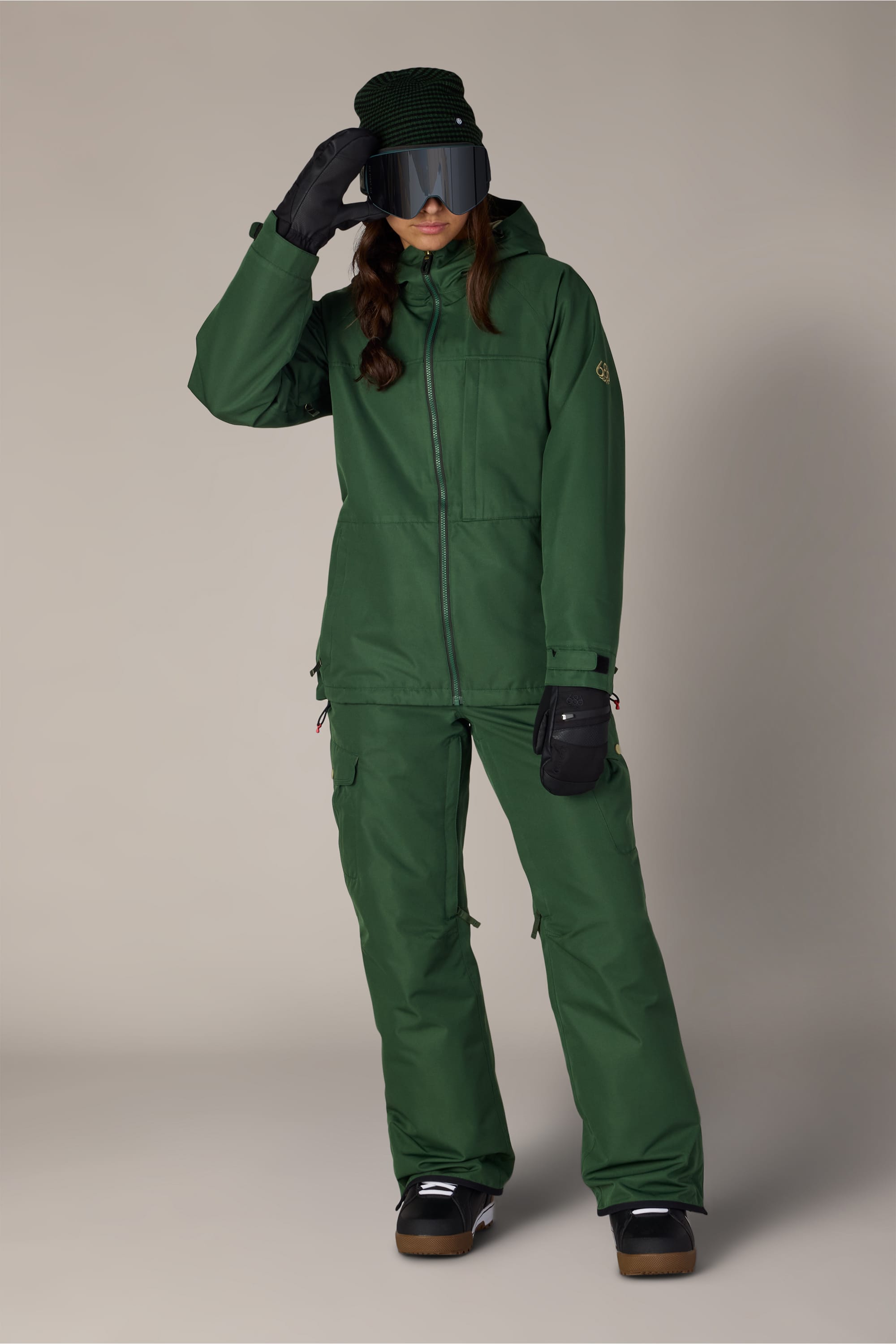 A person wearing the 686 Women’s Athena™ Insulated Jacket by 686, paired with matching green pants, black gloves, dark snow goggles, a green knit hat, and brown boots, posing against a plain background.