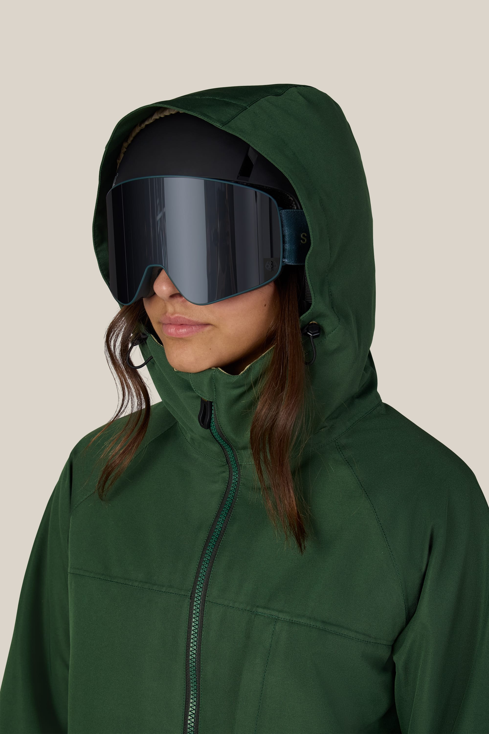 A person wearing the 686 Women’s Athena™ Insulated Jacket in green by 686, paired with large, reflective ski goggles and looking slightly to the side against a plain beige background.