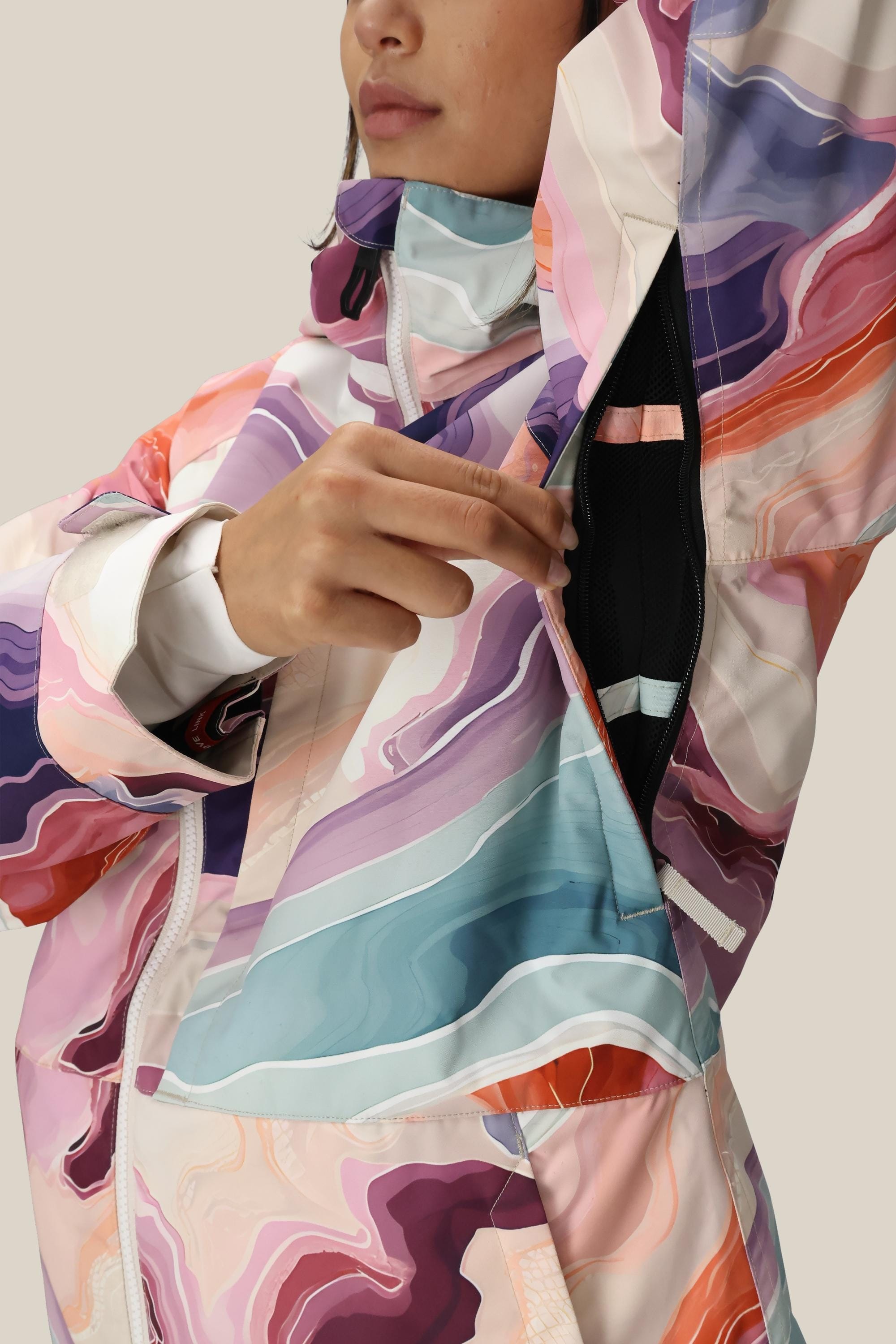 A woman in a 686 Women's Athena™ Insulated Jacket, featuring abstract swirls of pink, purple, blue, and beige, unzips the underarm vent. The waterproof jacket is by 686; her face is partially visible.