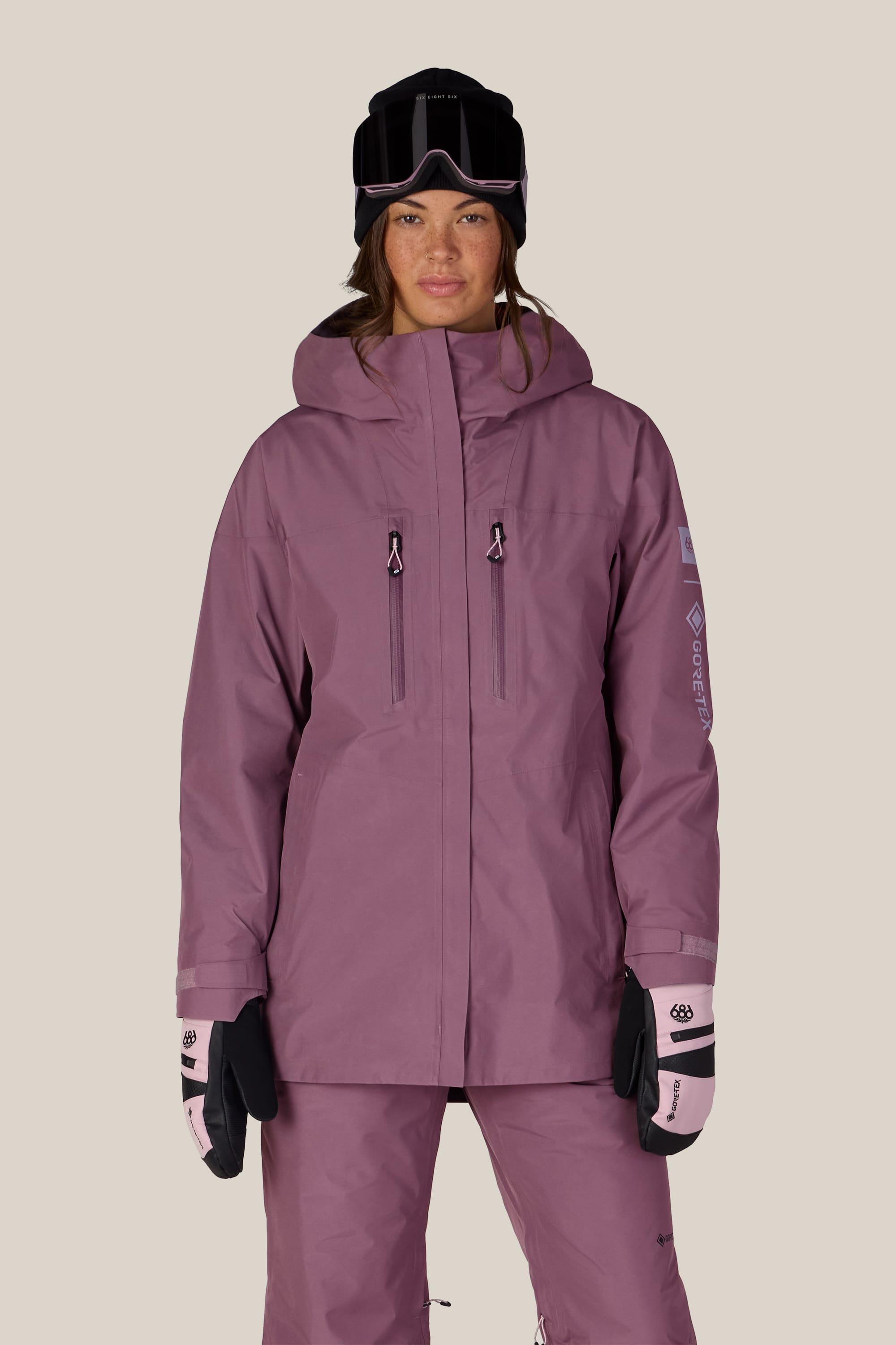 A person wearing the 686 Women’s GORE-TEX® Skyline Thermagraph® Jacket by 686 in mauve, with matching pants, a black beanie, ski goggles, and light pink mittens stands facing forward against a plain beige background.