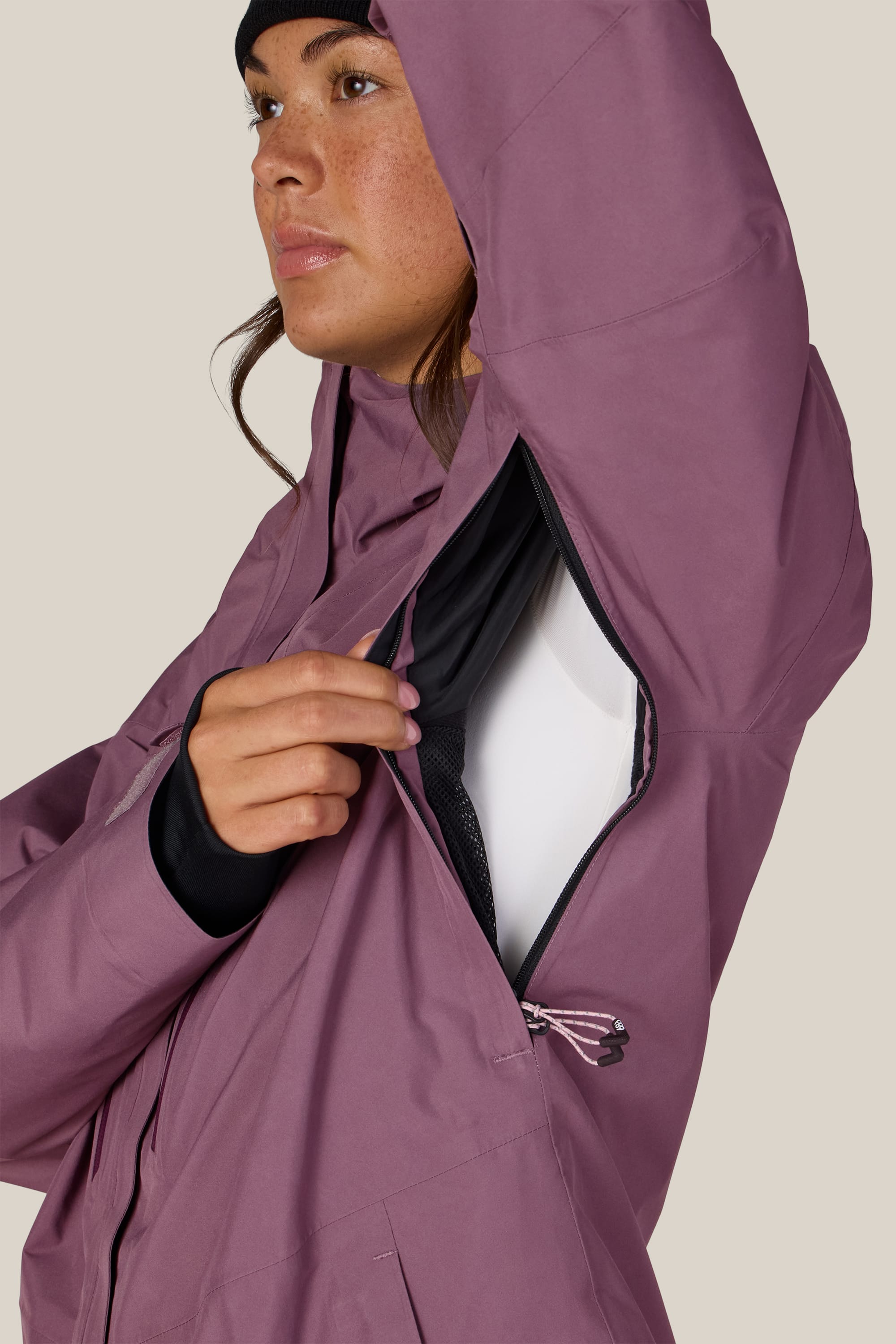 A person wearing the 686 Women’s GORE-TEX® Skyline Thermagraph® Jacket in mauve and a black beanie opens a zippered underarm vent, revealing the jacket’s black mesh lining. The background is plain and neutral.